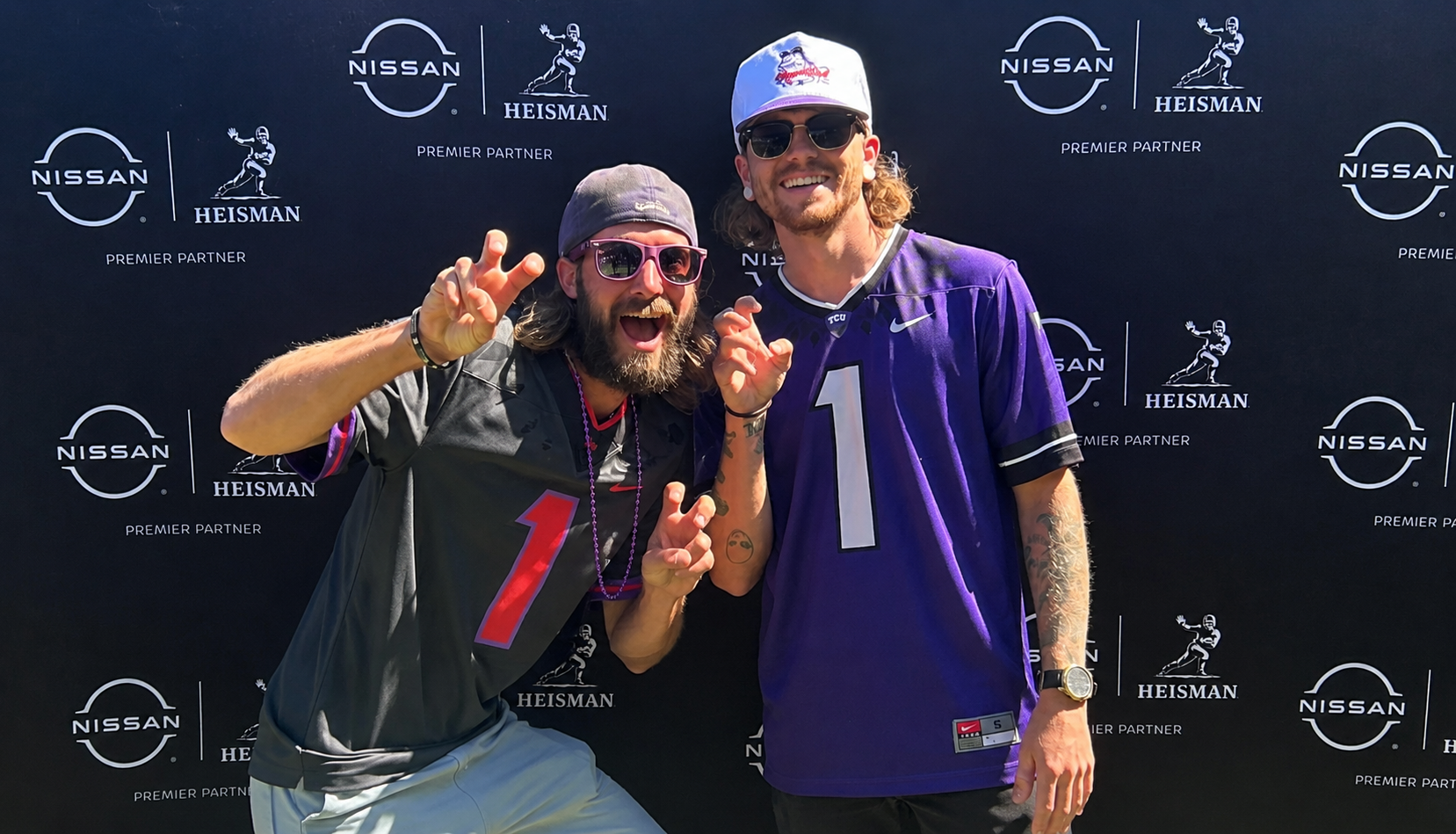 Two men posing in front of a black backdrop with Nissan and Heisman logos. The man on the left wears a black football jersey with the number 1, pink sunglasses, a gray cap, and is making a playful claw gesture. The man on the right wears a purple football jersey with the number 1, a white cap with a logo, sunglasses, and a watch, smiling while posing.