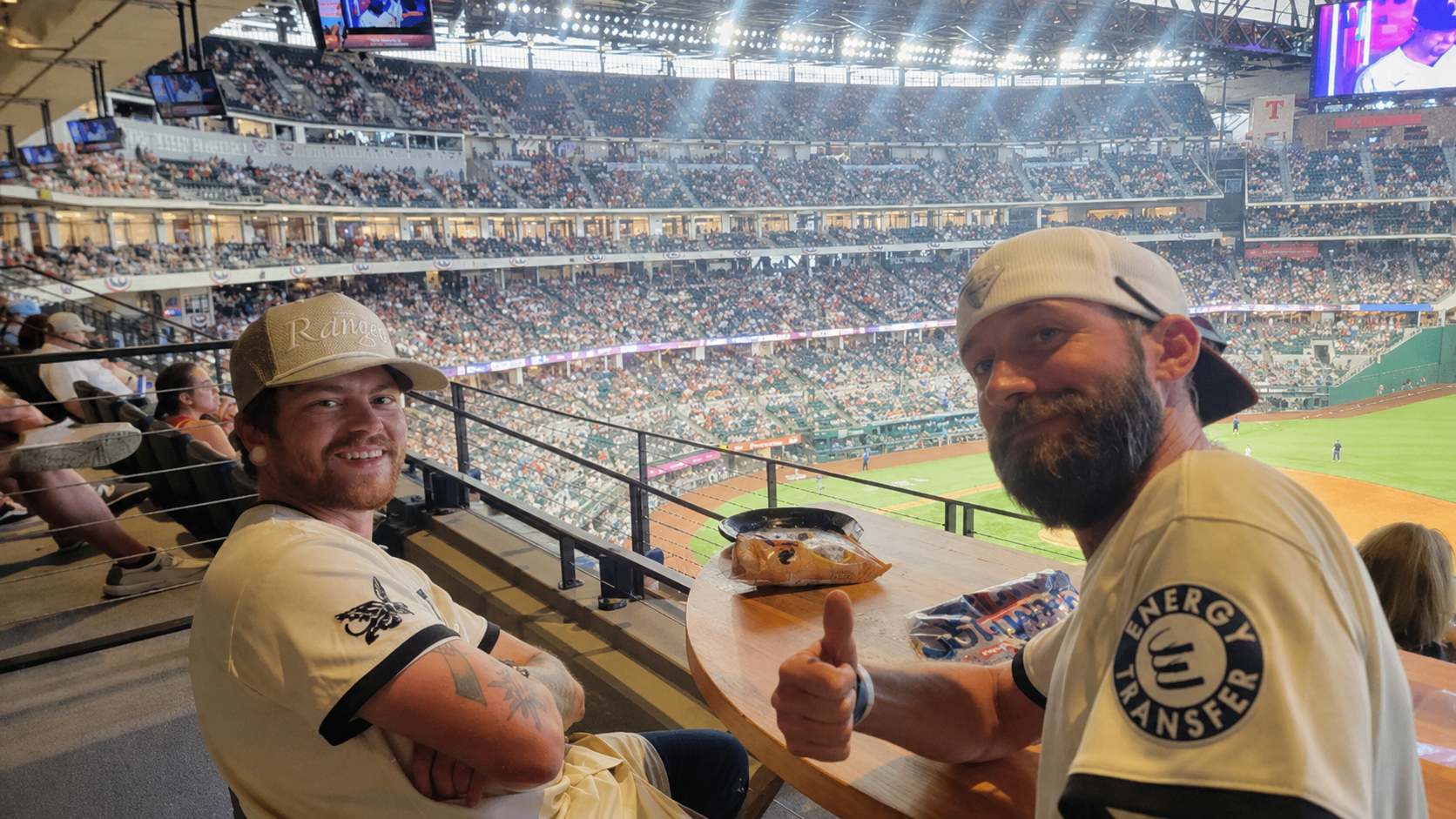 Two men sitting in the stands of a baseball stadium, smiling and giving a thumbs-up, with the baseball game visible in the background.