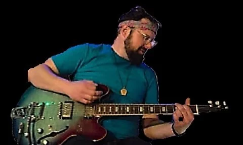 Person playing an electric guitar, wearing a turquoise shirt, glasses, and a cap, against a black background.