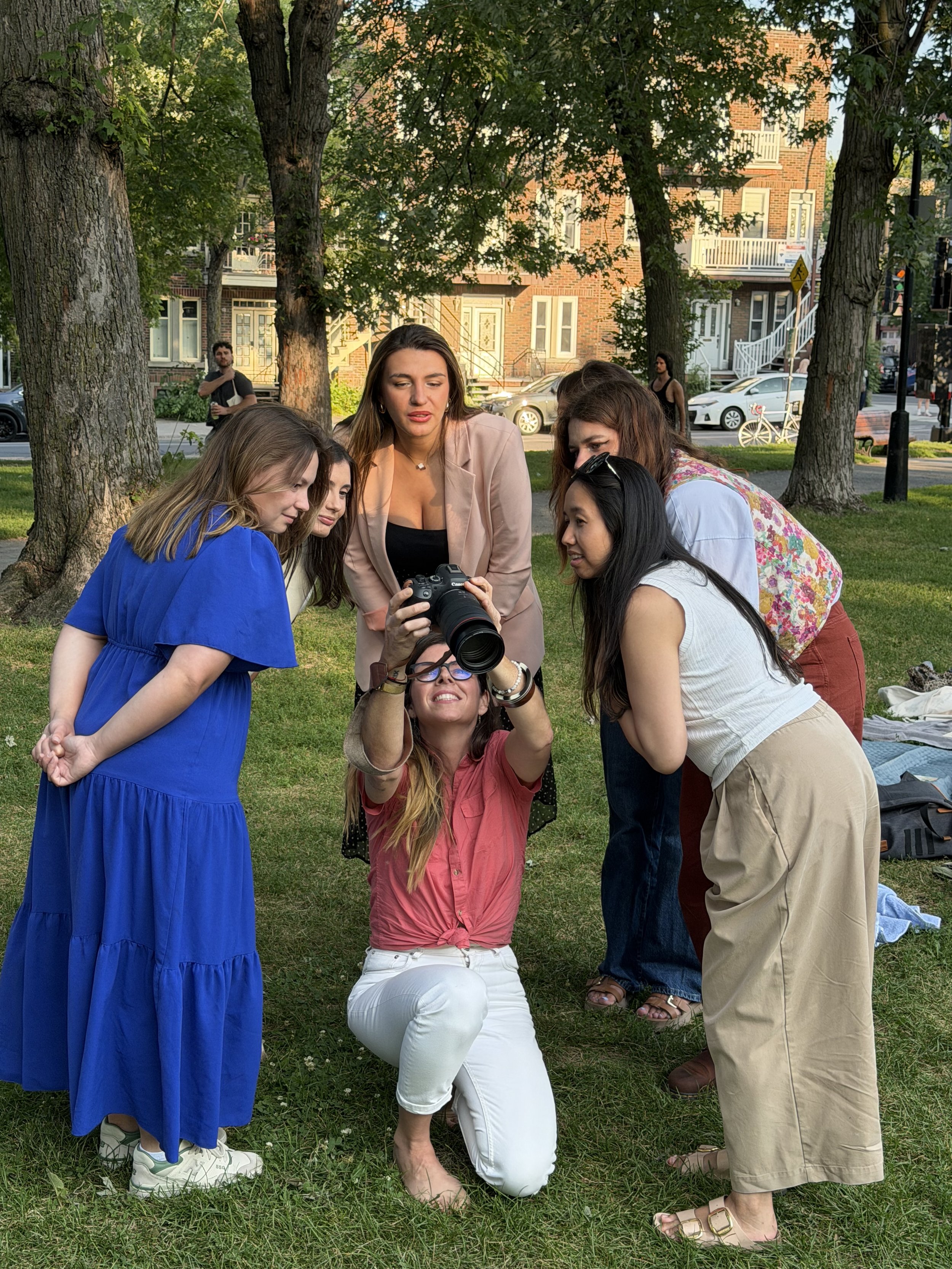 Un groupe de six femmes qui regardent une caméra numérique, dans un parc avec des arbres et des bâtiments résidentiels en arrière-plan.
