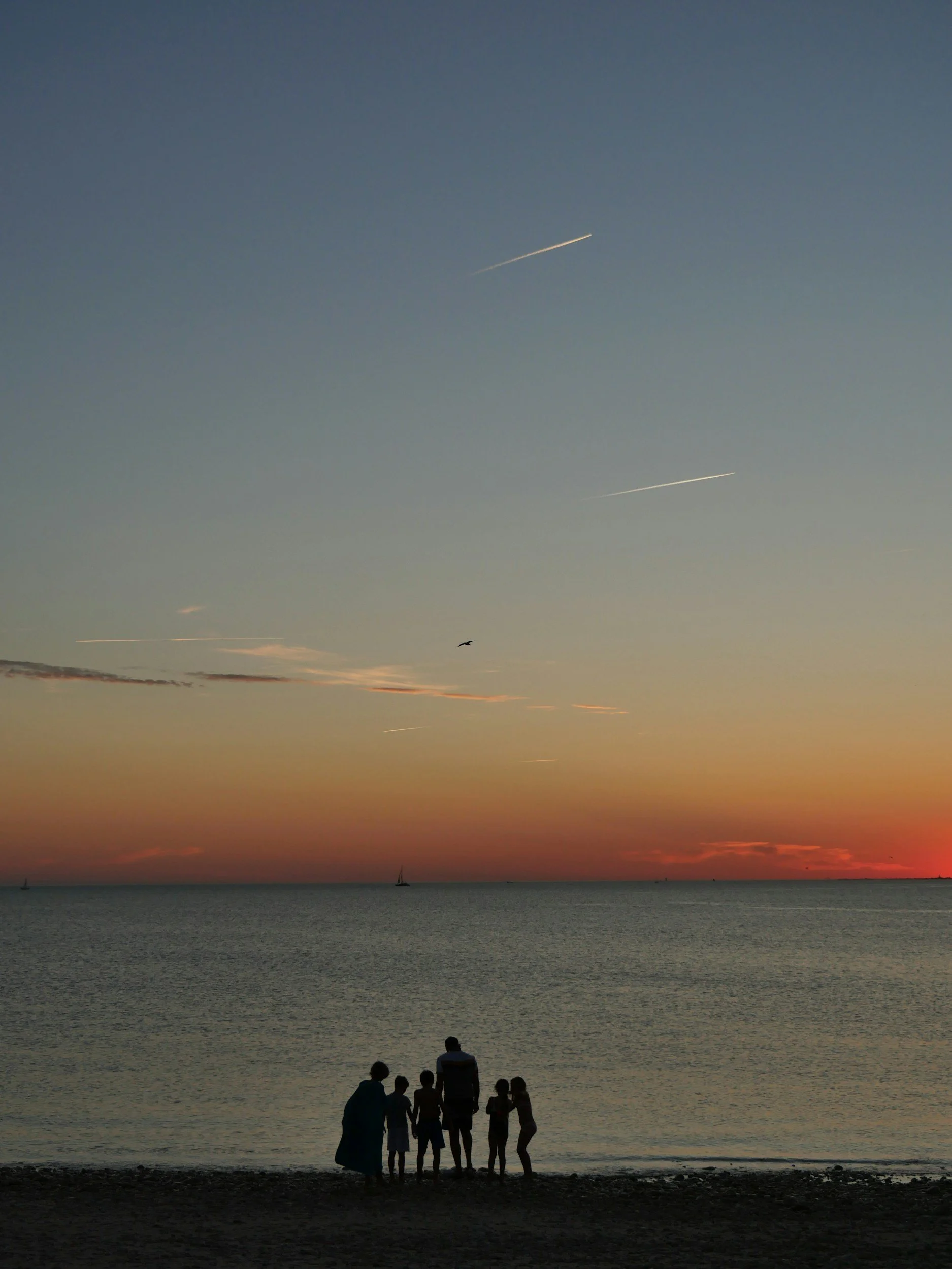 Groupe de personnes, dont des enfants et un adulte, debout sur la plage regardant la mer au coucher du soleil avec un ciel coloré et quelques avions laissant des traces dans le ciel.