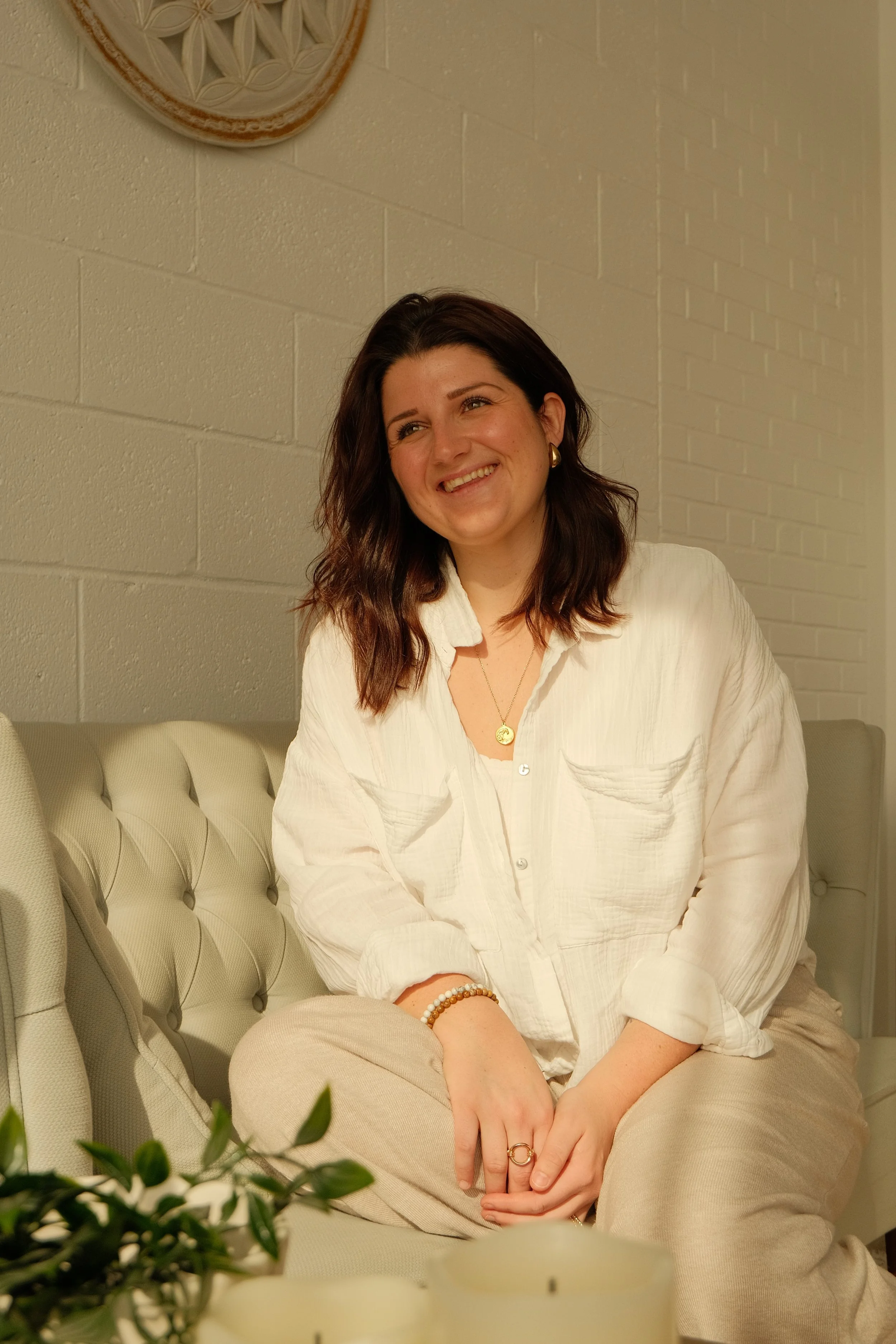 Une femme souriante assise sur un canapé beige, portant une chemise blanche et un pantalon beige. Elle porte des boucles d'oreilles dorées, un collier et un bracelet. La photo est prise en intérieur avec un mur en briques blanches en arrière-plan et une plante en premier plan.