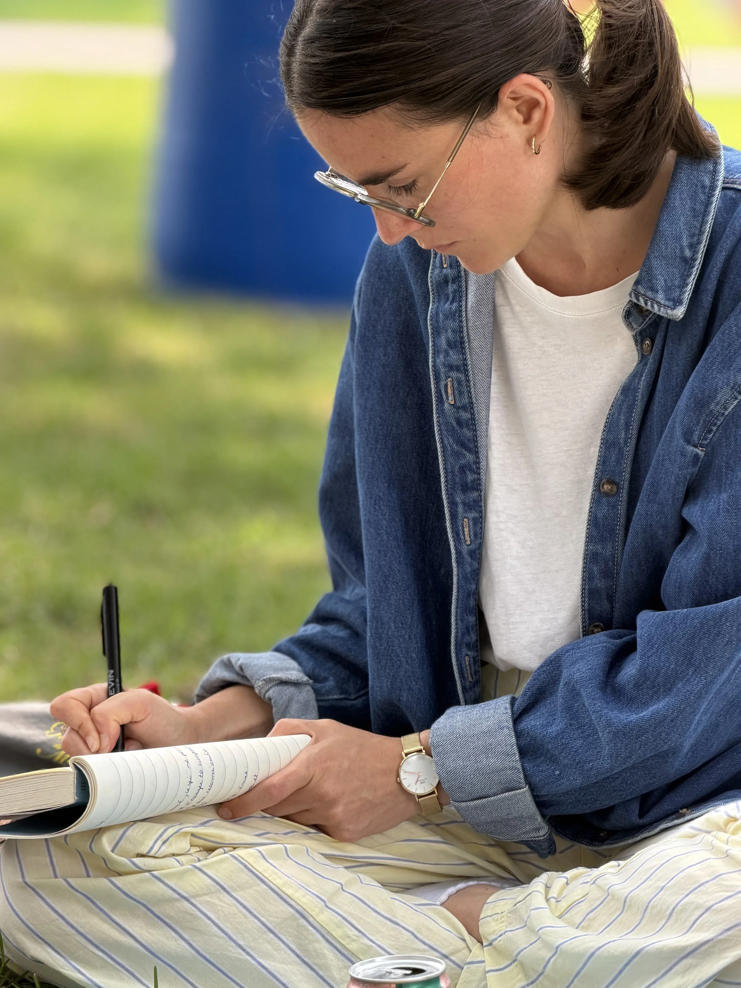 Une femme assise sur une couverture à rayures, écrivant dans un carnet avec un stylo, portant une montre dorée et des lunettes, avec un environnement extérieur vert en arrière-plan.