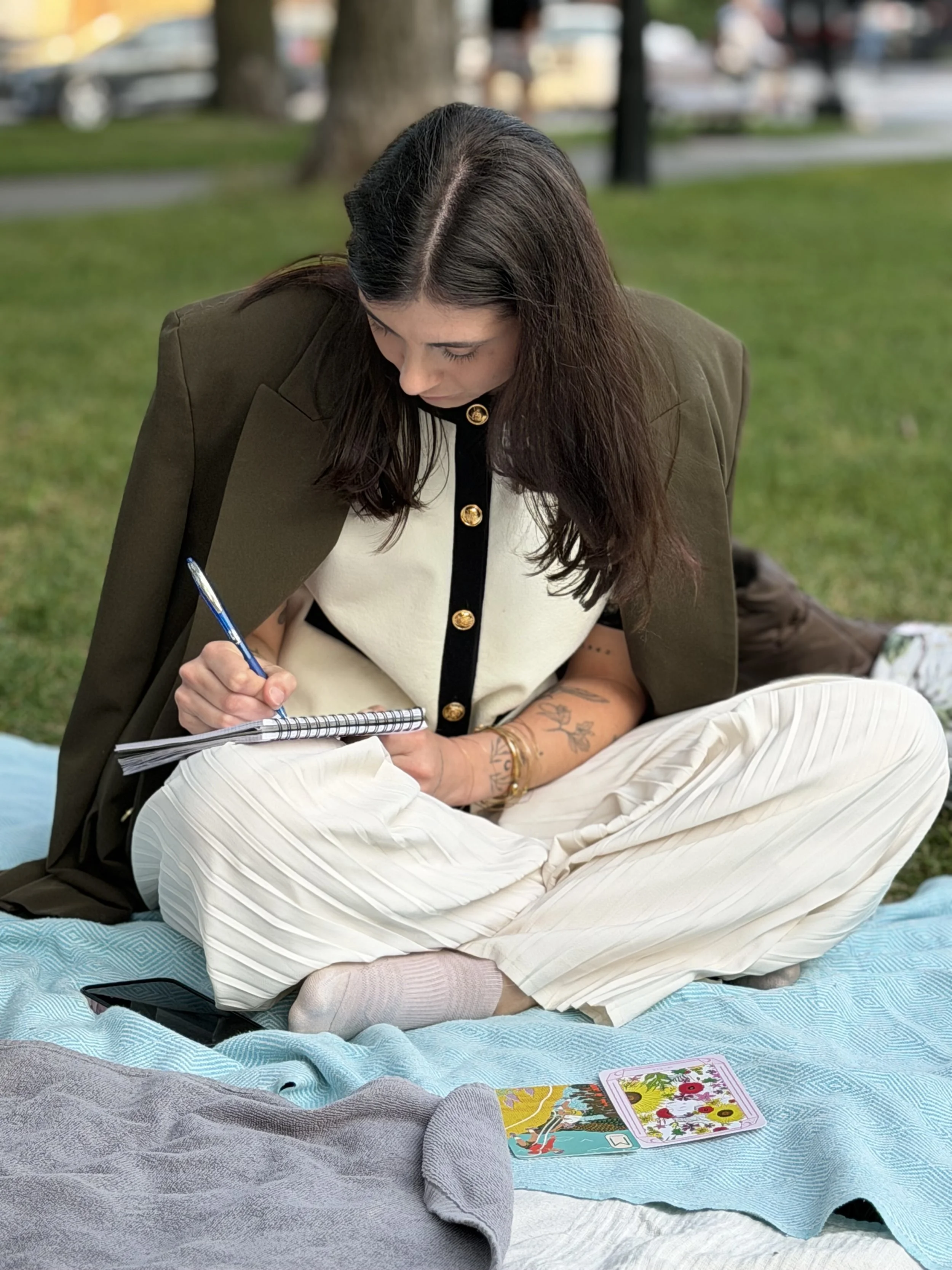 Une femme assise sur un tapis en plein air, en train d'écrire dans un carnet, avec des cartes de tarot posées devant elle.