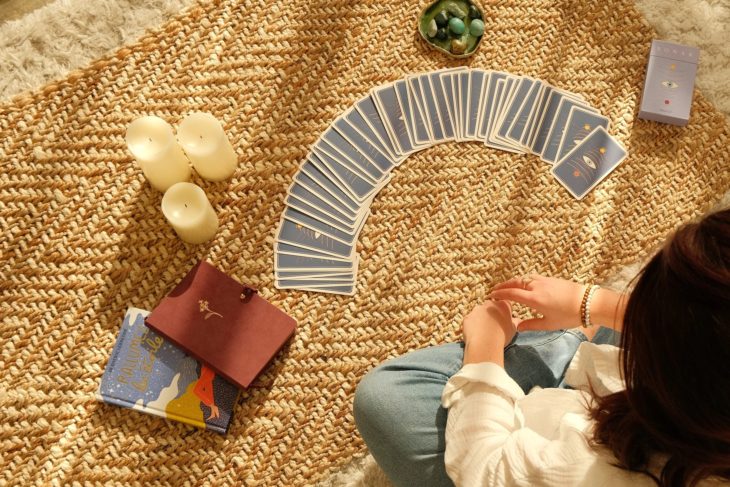 Jeune femme assise sur un tapis tressé, jouant à un jeu de tarot avec des cartes bleu foncé, entourée de trois bougies blanches, un petit plateau avec des pierres semi-précieuses, un livre, une boîte de cartes oracle et d'autres objets.