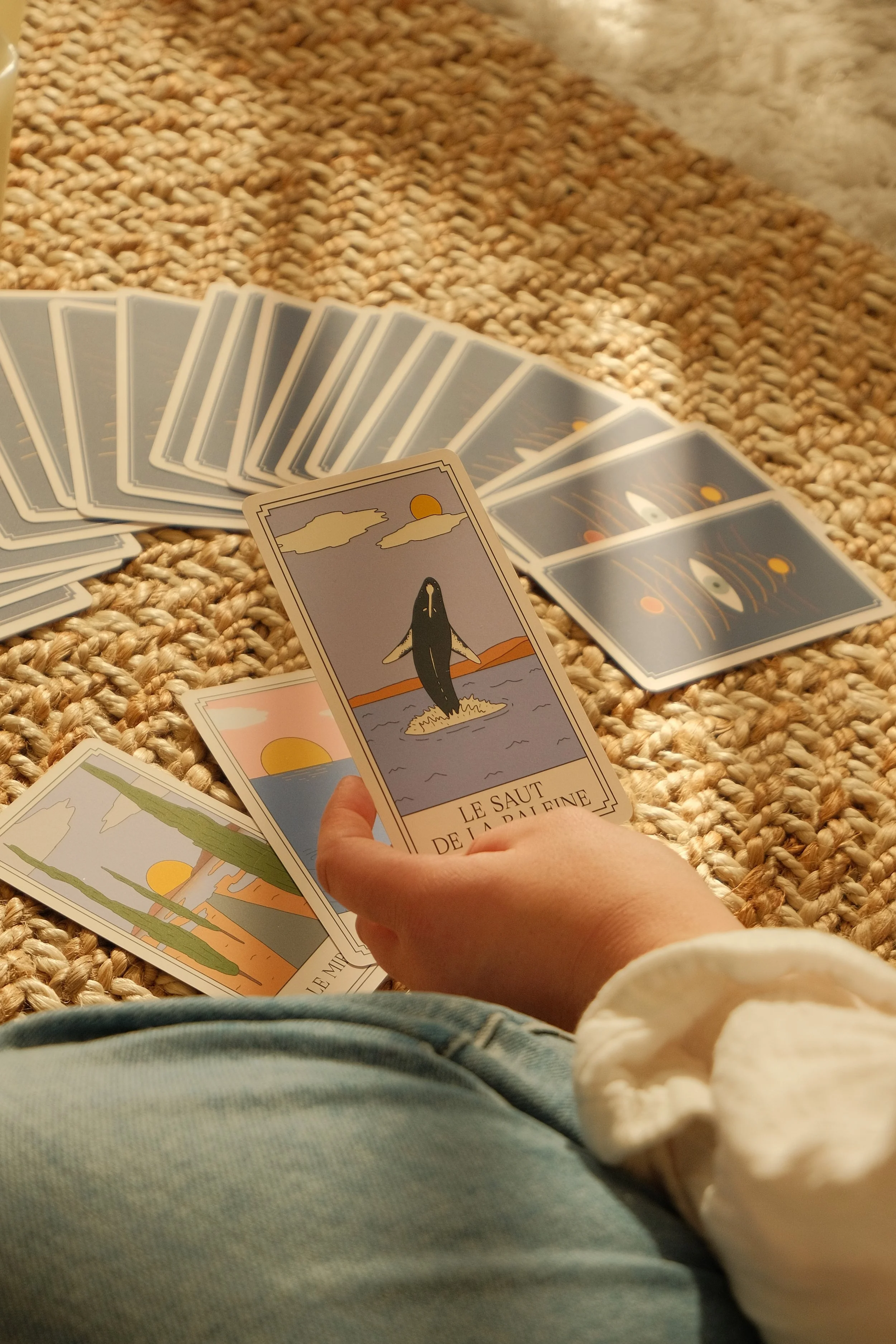 Mains tenant un jeu de cartes de tarot sur un tapis en laine, avec d'autres cartes éparpillées autour. La carte visible montre un dauphin sautant hors de l'eau au coucher du soleil.