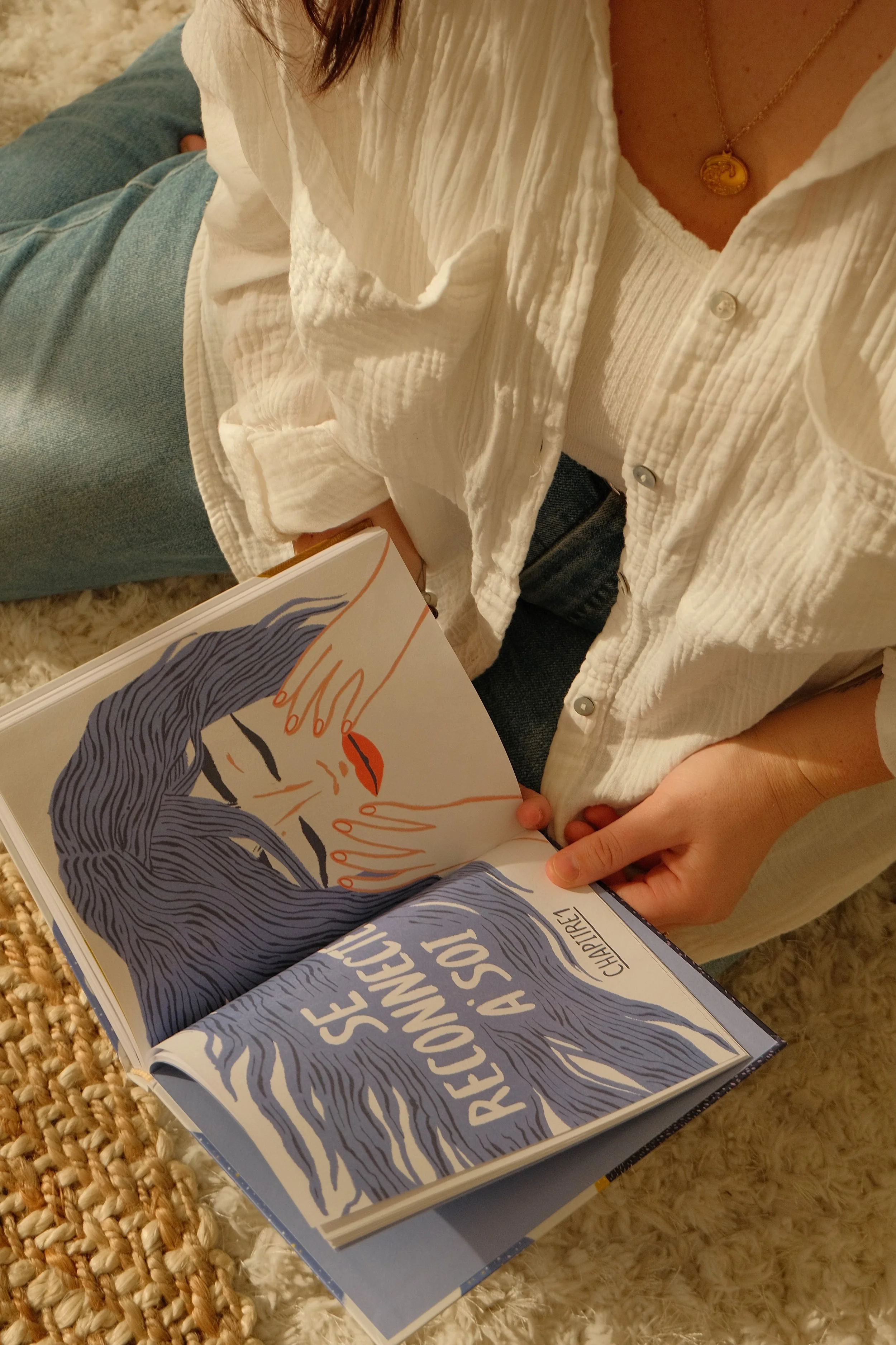 Une personne tient un livre ouvert qui montre une illustration d'une femme avec des cheveux longs, portant des lunettes, avec les mains sur le visage. La personne porte un chemisier blanc et une chaîne avec un pendentif.