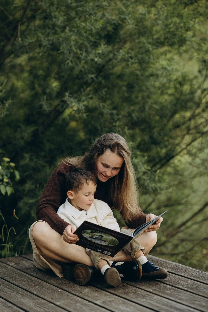 Eine Mutter mit ihrem Sohn sitzen auf einem Holzdeck in der Natur, lesen gemeinsam ein Buch und genießen die Zeit während eines entspannten Familienshootings in Klagenfurt, Kärnten.