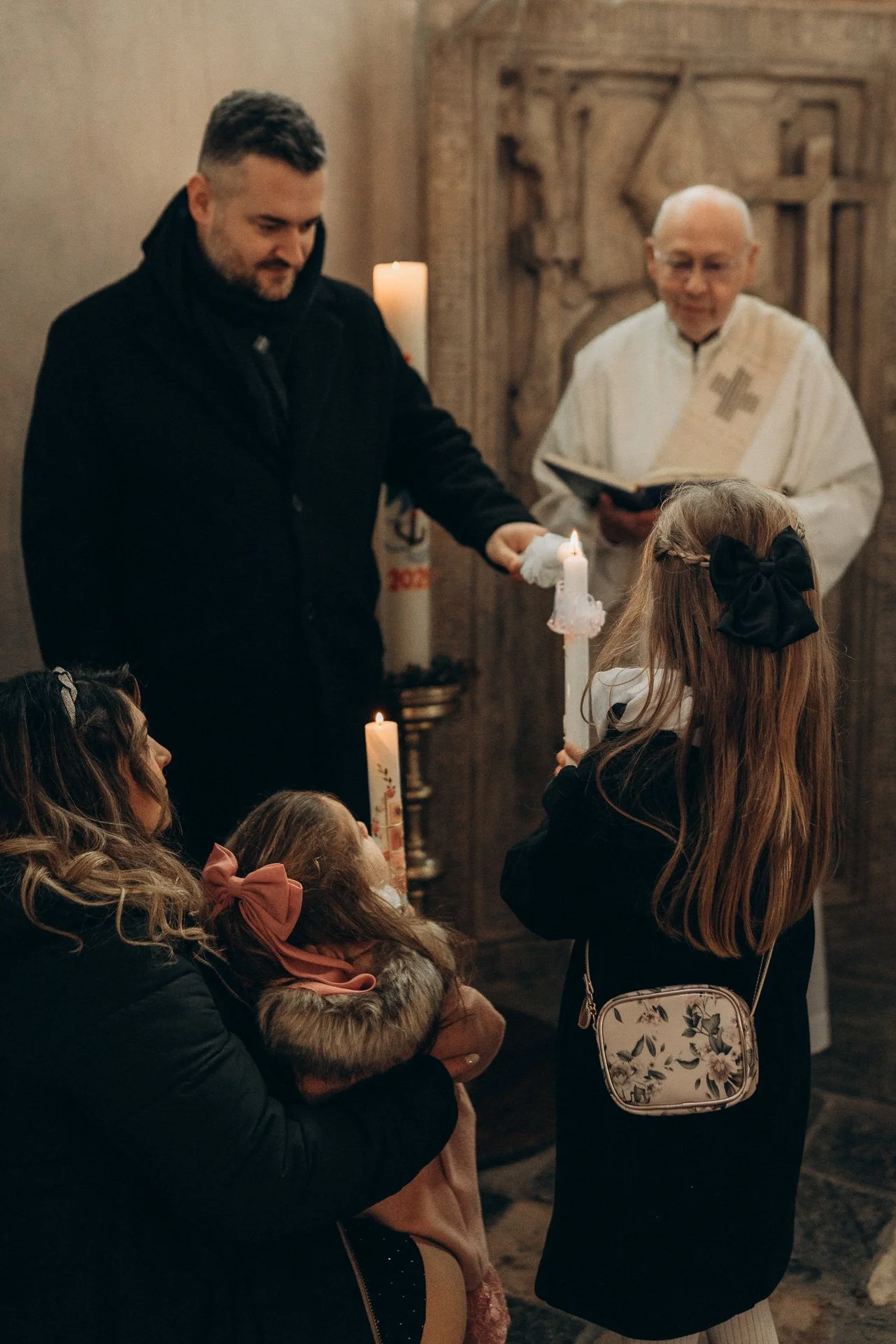 Ein Mann mit dunkler Jacke übergibt einer jungen Frau mit langem Haar und schwarzem Kleid eine brennende Kerze in einer Kirche, im Hintergrund ein Priester bei der Lesung, umgeben von mehreren Kindern und Erwachsenen.