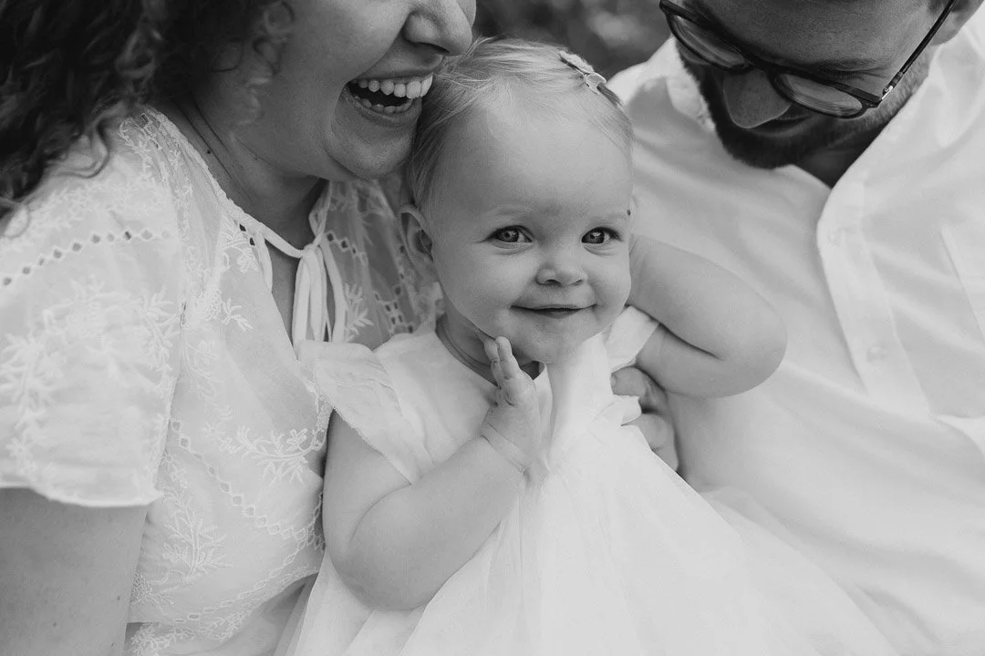 Ein kleines, lachendes Mädchen, umgeben von seinen Eltern bei einem Familienfotoshooting im Freien in Klagenfurt, Kärnten.