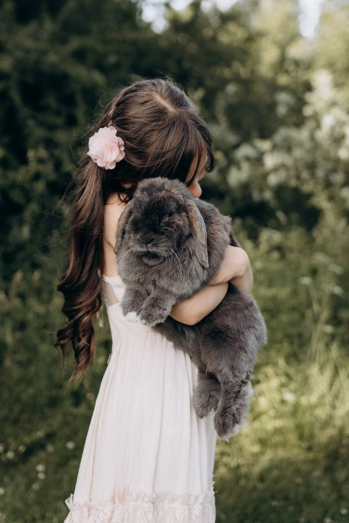 Ein Mädchen in einem weißen Kleid hält einen großen, grauen Hasen im Freien mit grüner Vegetation im Hintergrund.
