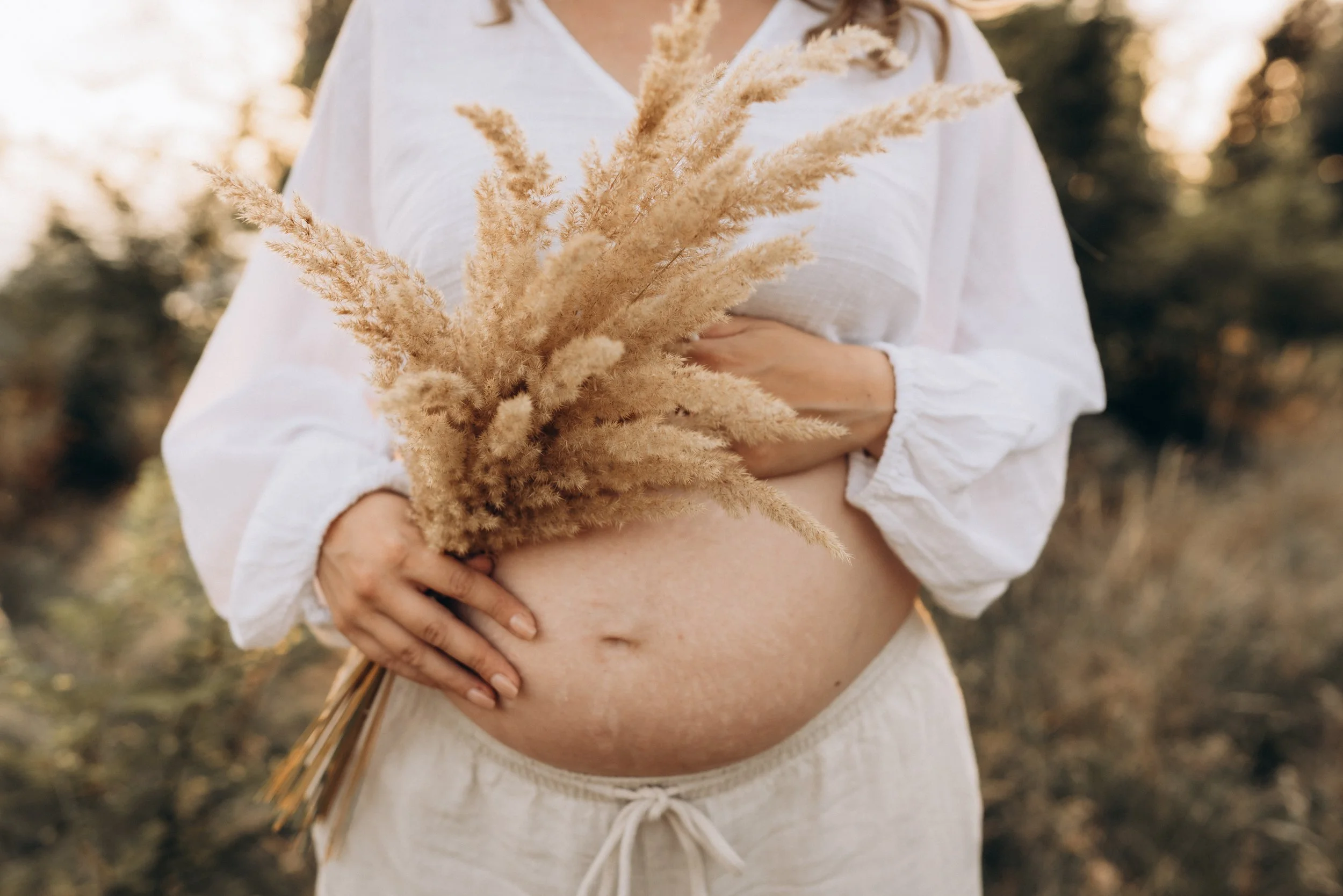 Schwangere Frau mit Trockengräsern im warmen Licht des Sonnenuntergangs beim Babybauch-Fotoshooting in Klagenfurt, Kärnten