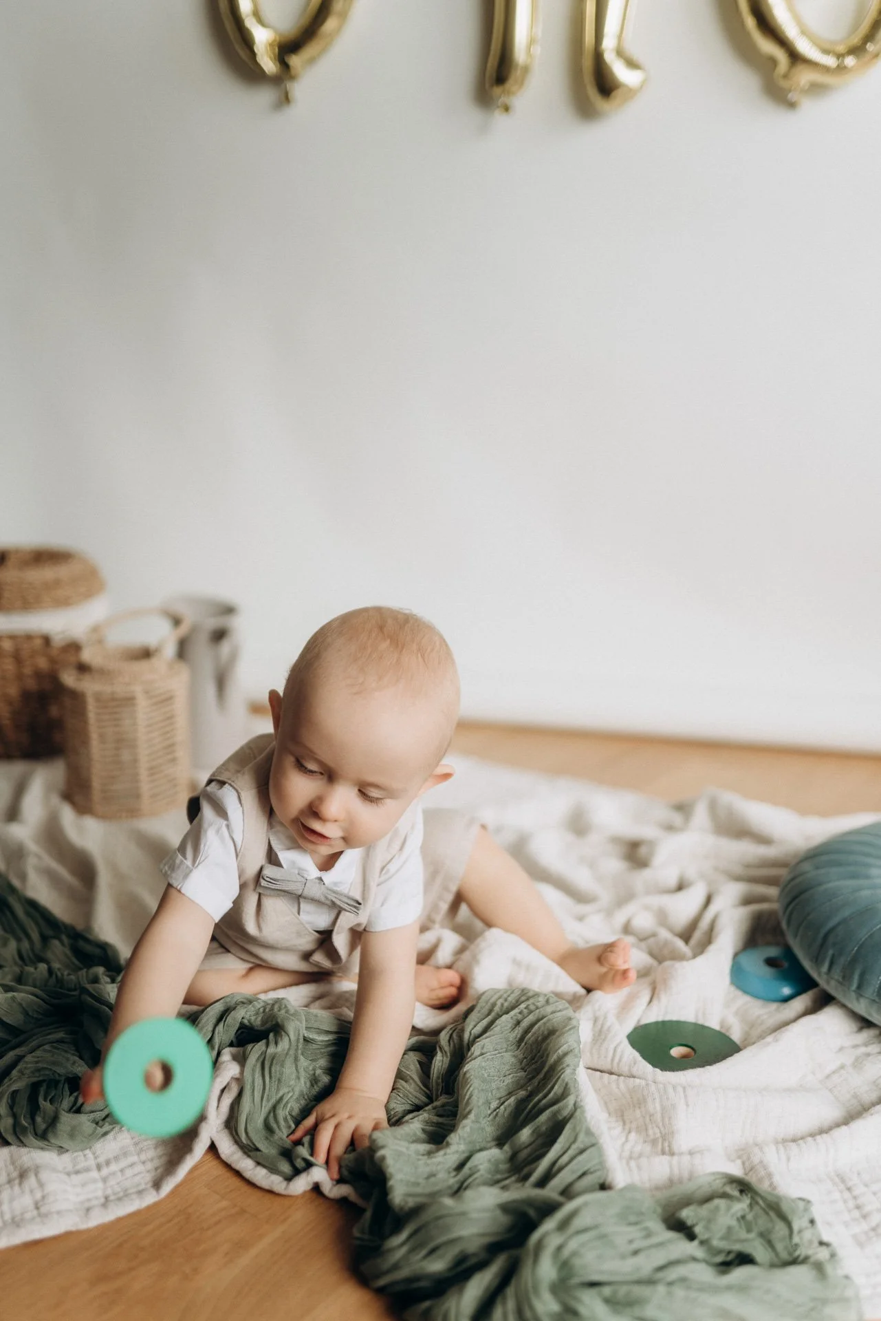 Ein Baby spielt auf einem Bett mit Decken und Kissen, neben Wäschekörben und Spielsachen, in einem hellen Raum.