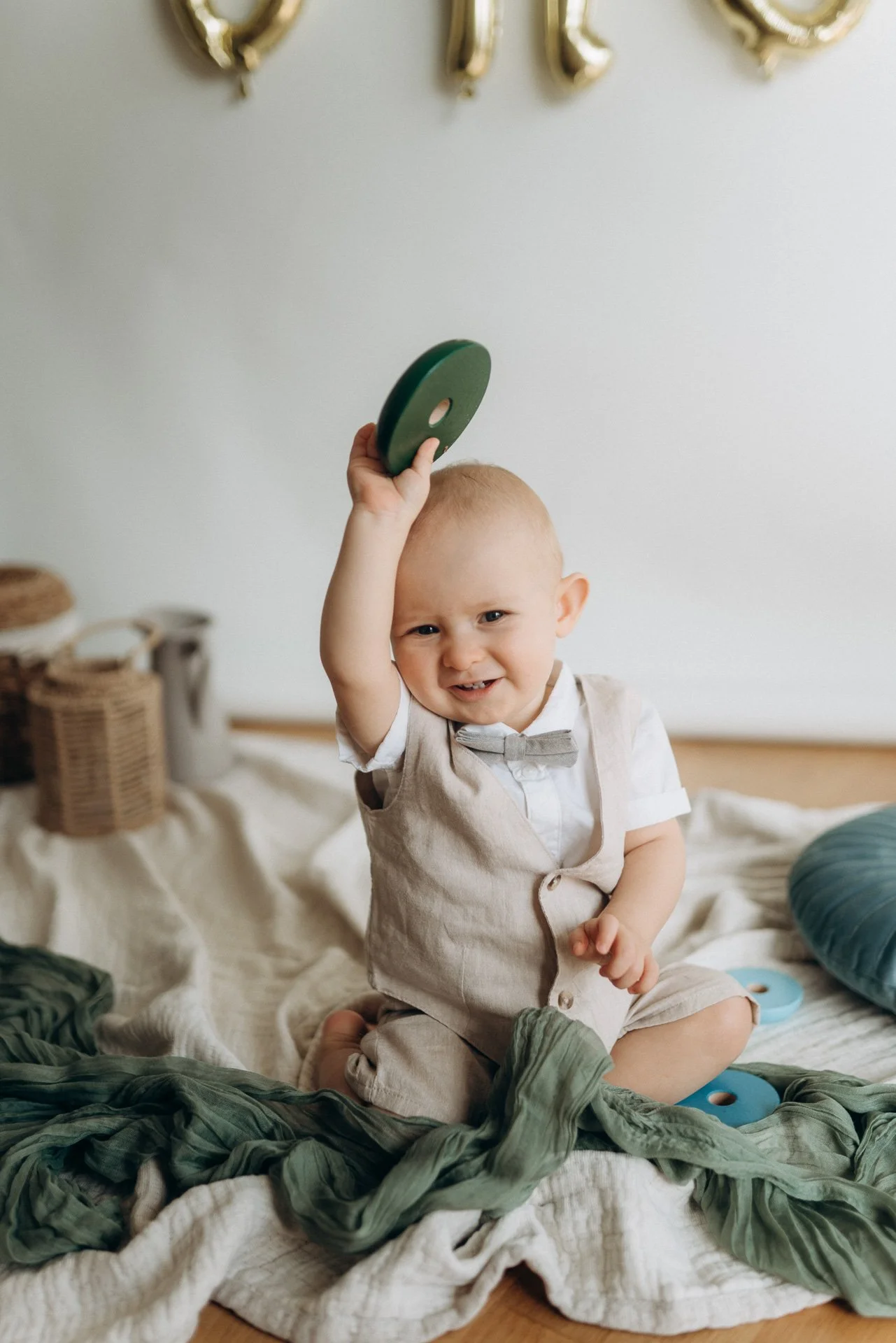 Ein lachendes Baby in einem hellen Raum, das ein grünes Spielzeug hält, auf einem Bett mit weichen Decken und Kissen, im Hintergrund Körbe und Dekorationen.