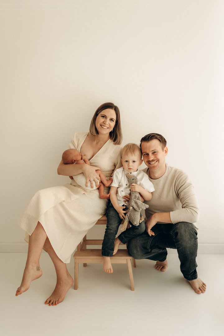 Familie mit zwei Kindern, eines ist ein Baby, sitzend und im weißen Kleid, das andere ein Kleinkind mit weißen Hemd und schwarzen Hosen, sitzt auf einem kleinen Stuhl, vor einem weißen Hintergrund.