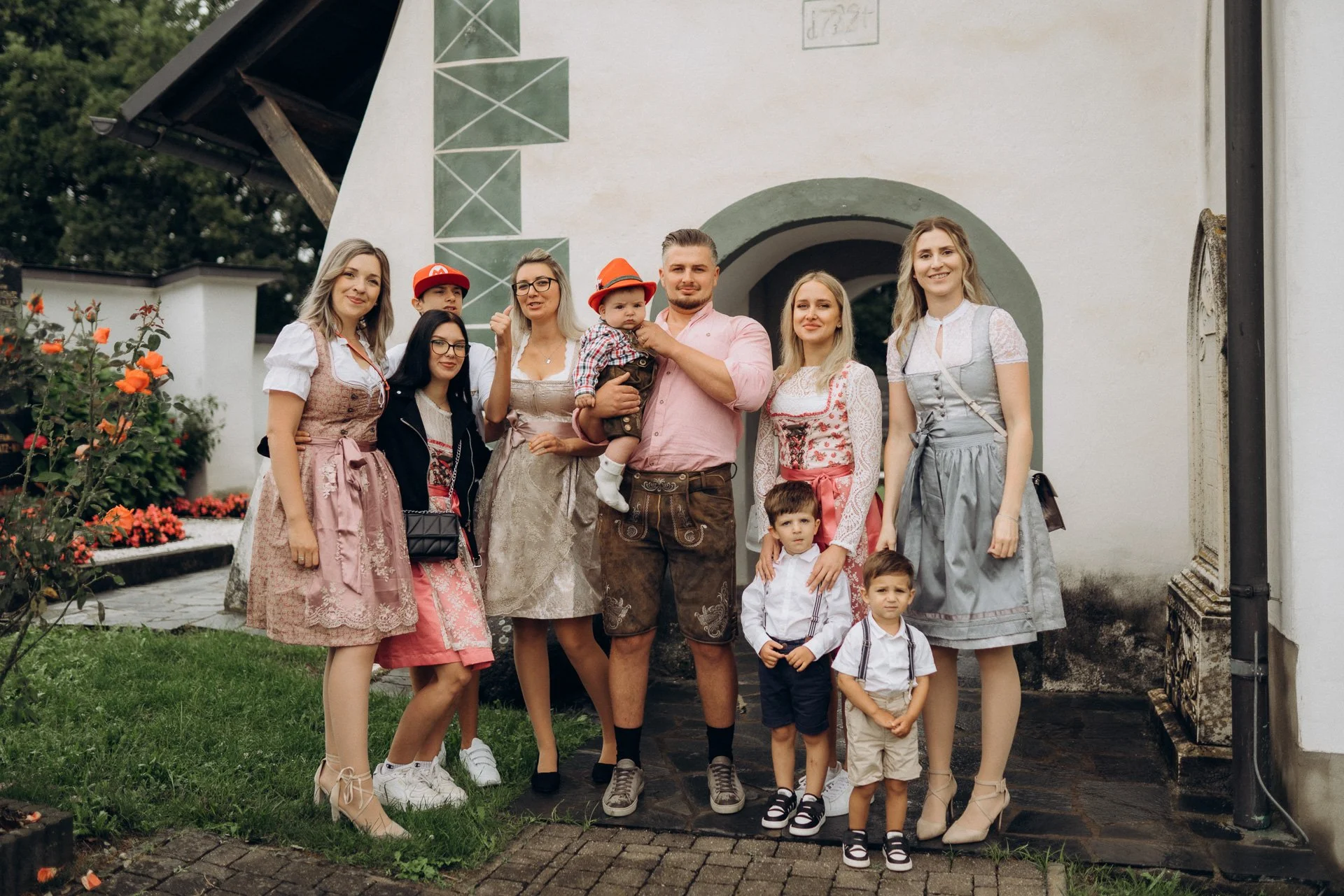 Eine Gruppe von Erwachsenen und Kindern in traditionellen dirndl- und lederhosenartigen Kleidern vor einem weißen Gebäude mit einem Bogen und grünen Dekorationen.
