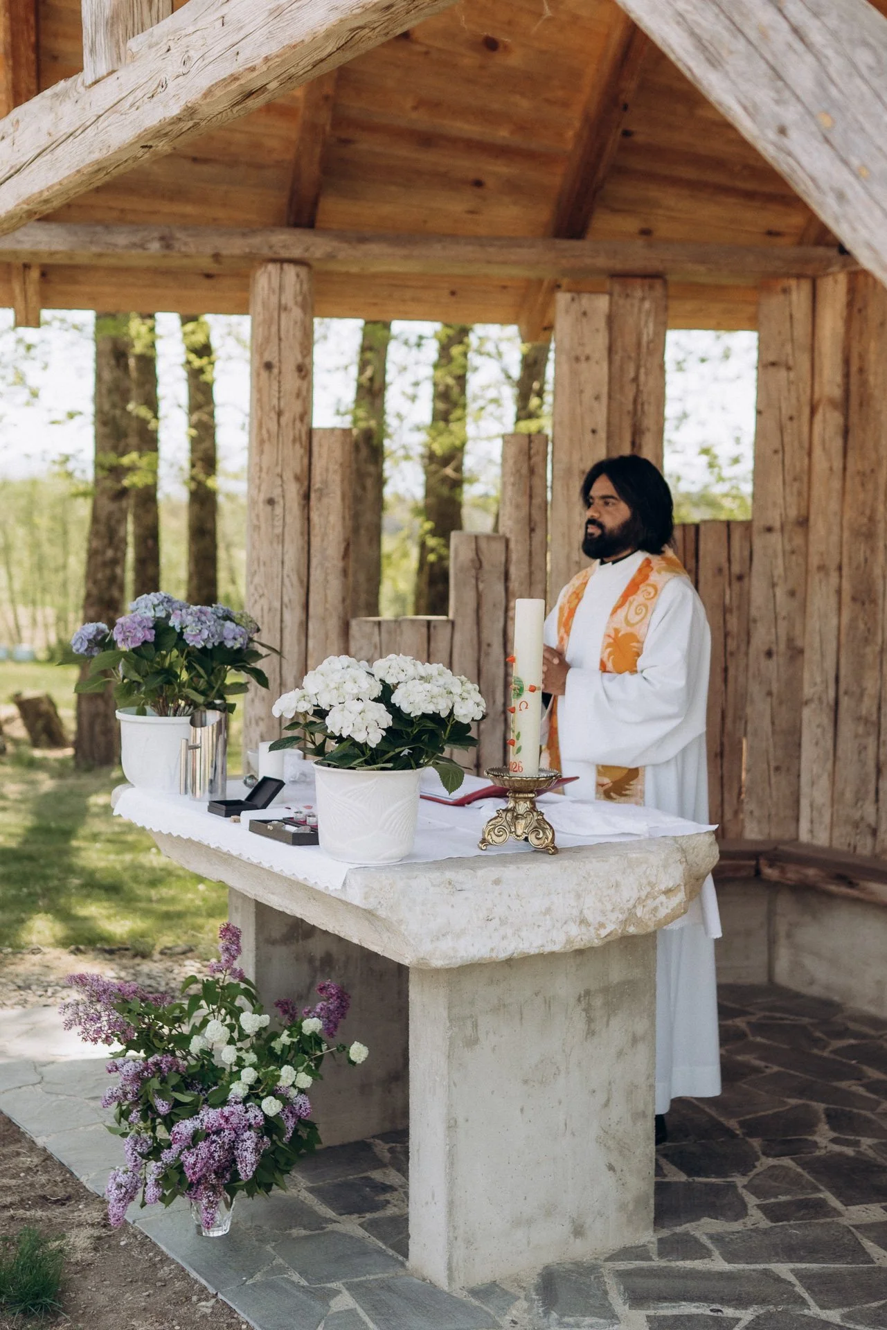 Ein Priester steht an einem Altar im Freien, umgeben von Blumen und Holzstrukturen, bei einer kirchlichen Zeremonie.