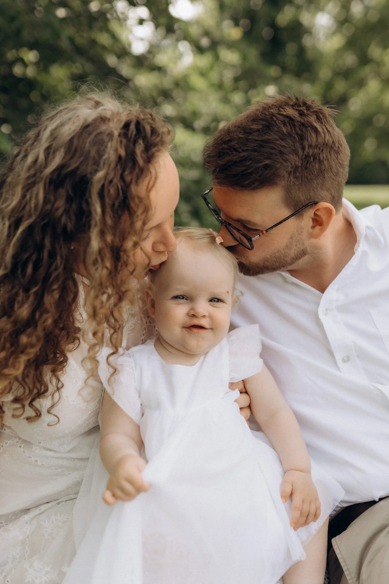 Familie mit einem kleinen Baby, das im Freien bei Bäumen sitzt. Der Vater und die Mutter küssen das Baby auf den Kopf.
