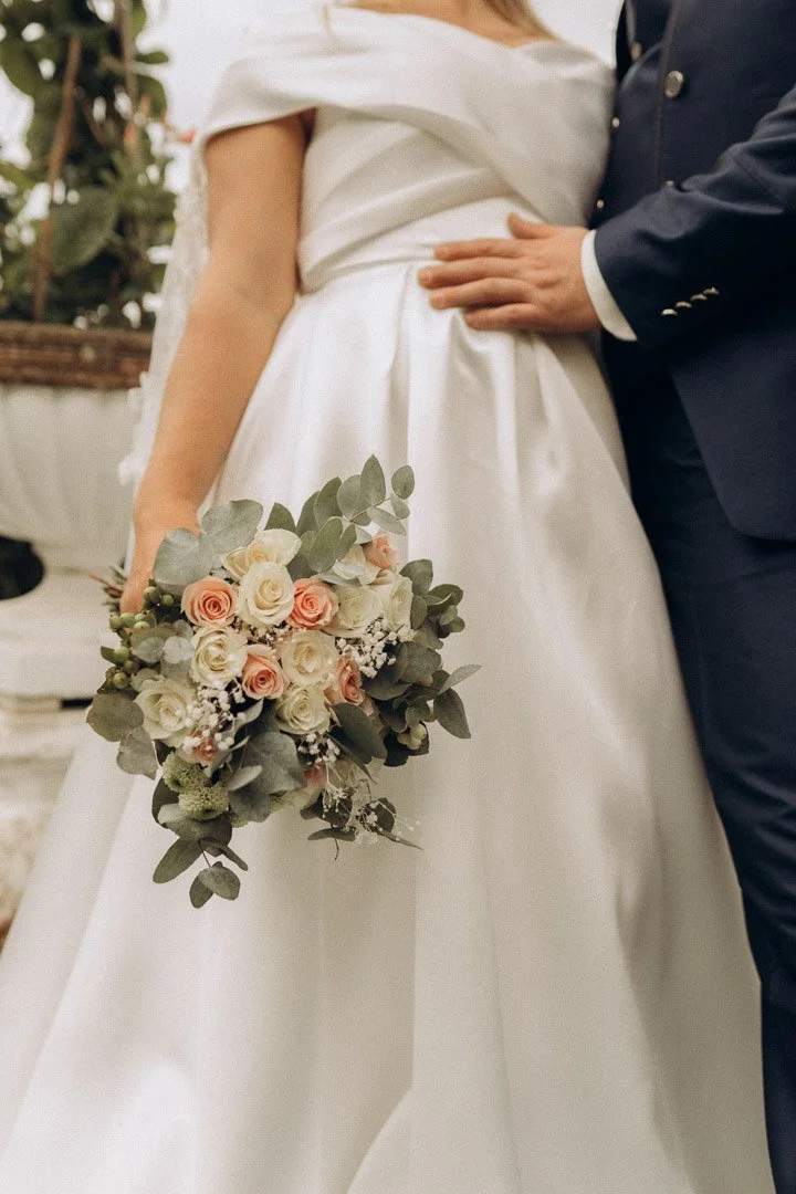 Braut mit weißem Brautkleid hält einen Blumenstrauß aus roses, Eukalyptus und anderen Blumen, während der Bräutigam in dunklem Anzug neben ihr steht.