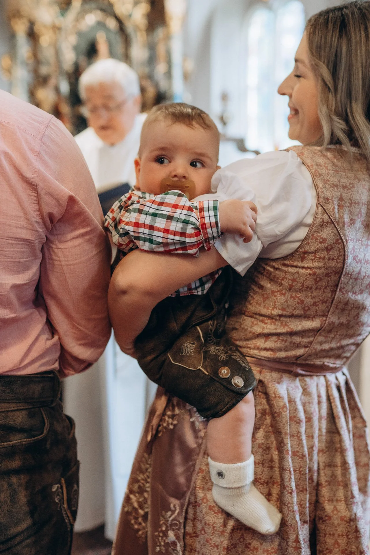 Ein Baby mit Schnuller wird von einer Frau in traditionellen Trachten gehalten, während im Hintergrund eine älterer Mann in ähnlicher Tracht zu sehen ist.