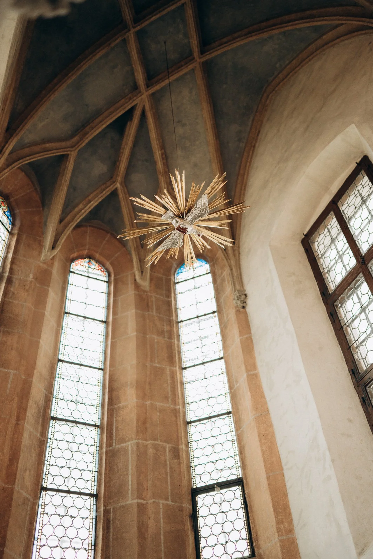 Ein Blick nach oben auf die Fenster und die gewölbte Decke eines Kirchenraums mit einem modernen, stilisierten Kreuzleuchter in der Mitte.