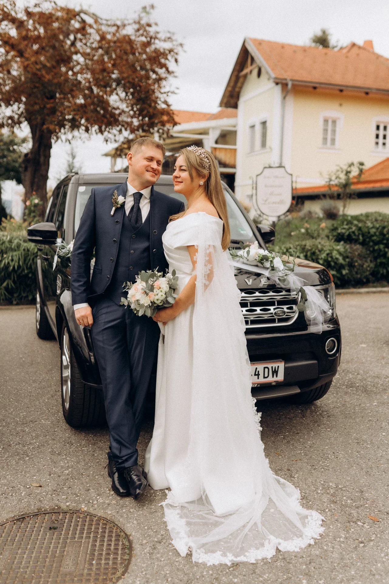 Ein glückliches Brautpaar posiert bei der Hochzeit vor einem schwarzen, mit weißen Blumen und Bändern dekorierten SUV als Hochzeitsauto bei einem Hochzeitsshooting in Klagenfurt.