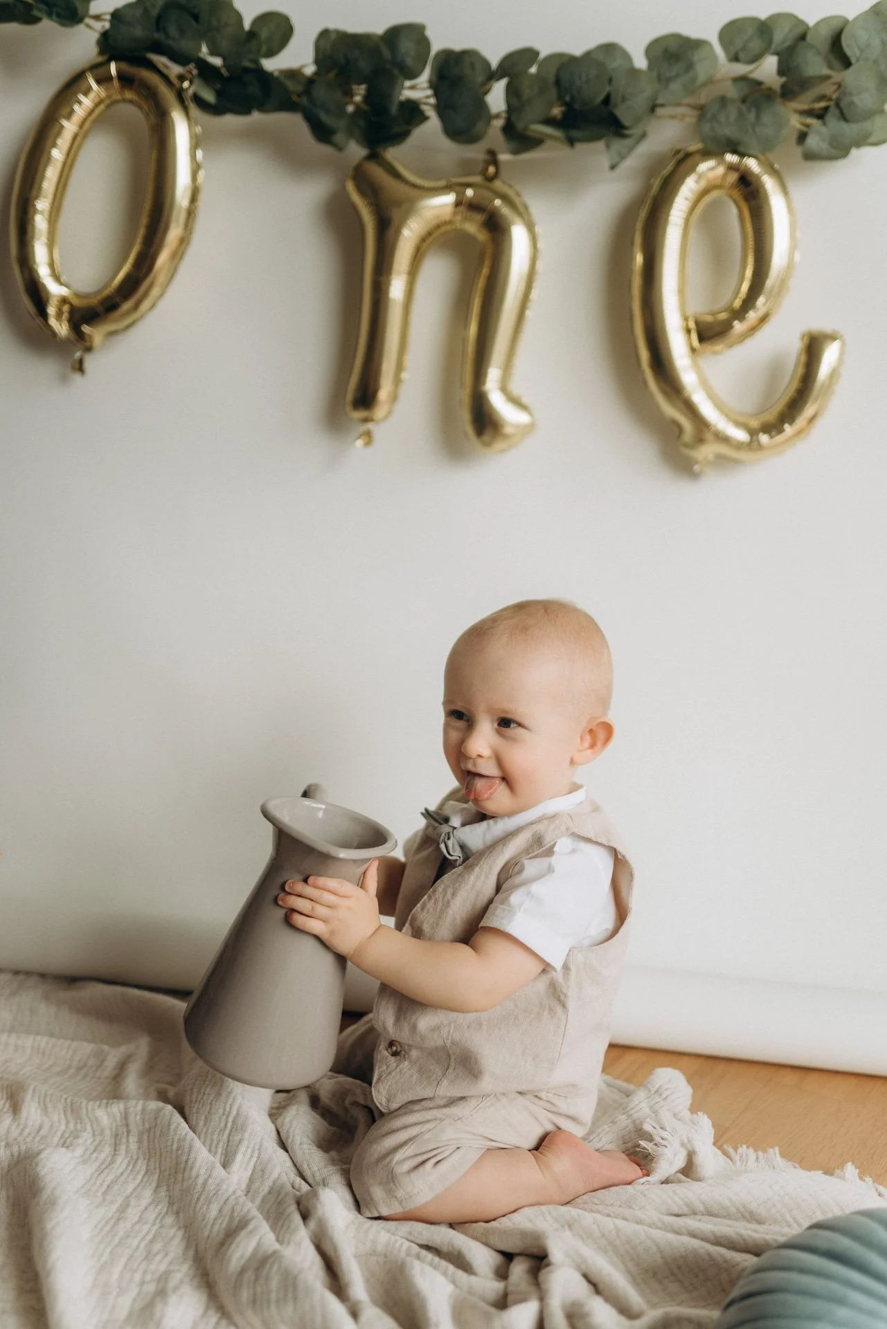 Ein kleiner Junge kniet auf einem weichen, cremefarbenen Bettuch, hält eine große graue Vase mit beiden Händen und schaut lachend nach oben. Über ihm hängen goldene Ballons, die die Buchstaben 'ONE' bilden, und grüne Zweige schmücken das obere Bild.