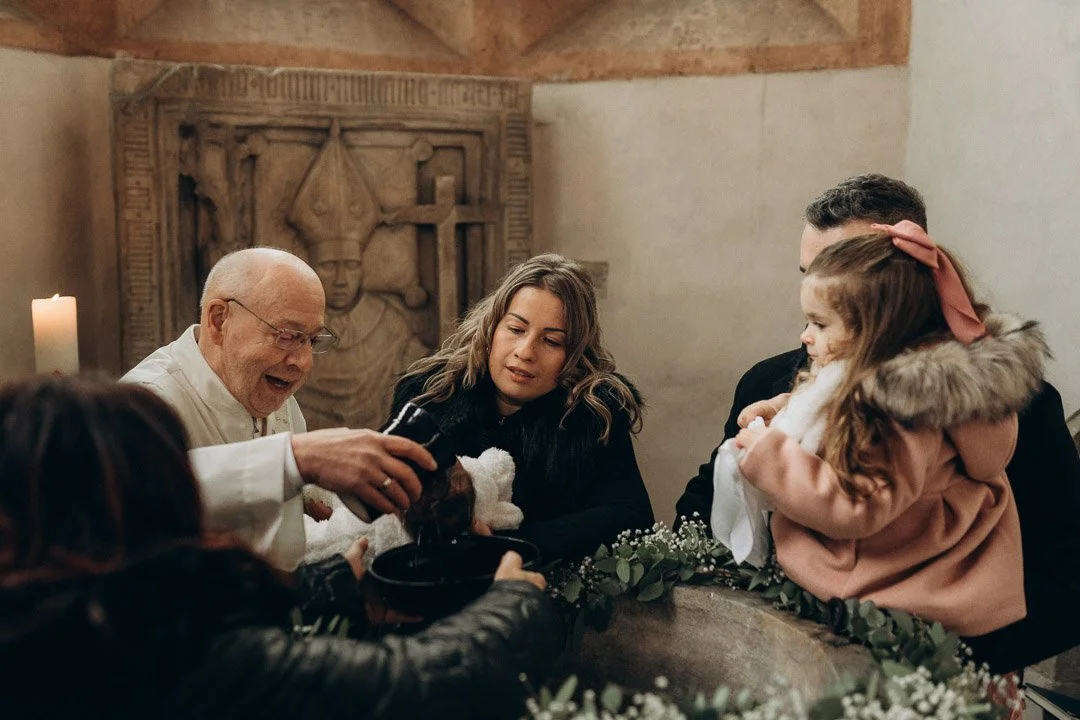Familie bei einer Tauferinnerung in einer Kirche, mit einem Priester und eine Taufe mit Wasser, Eltern und Kind sind anwesend.