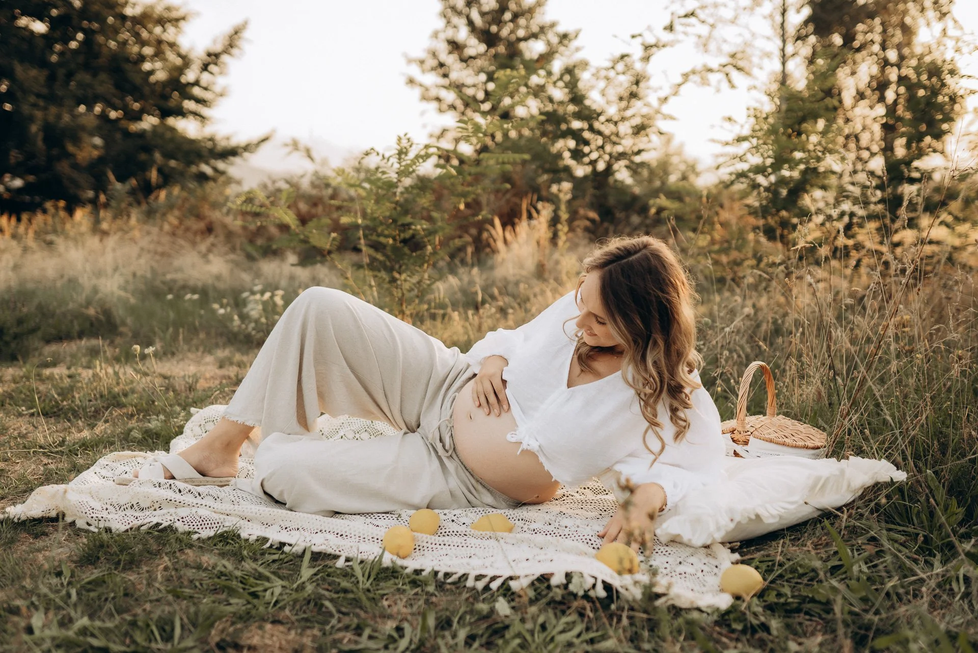 Schwangere Frau liegt auf einer Decke im Freien, umgeben von Bäumen, mit Sonnenlicht im Hintergrund, hält ihren Bauch, während sie lächelnd schaut.