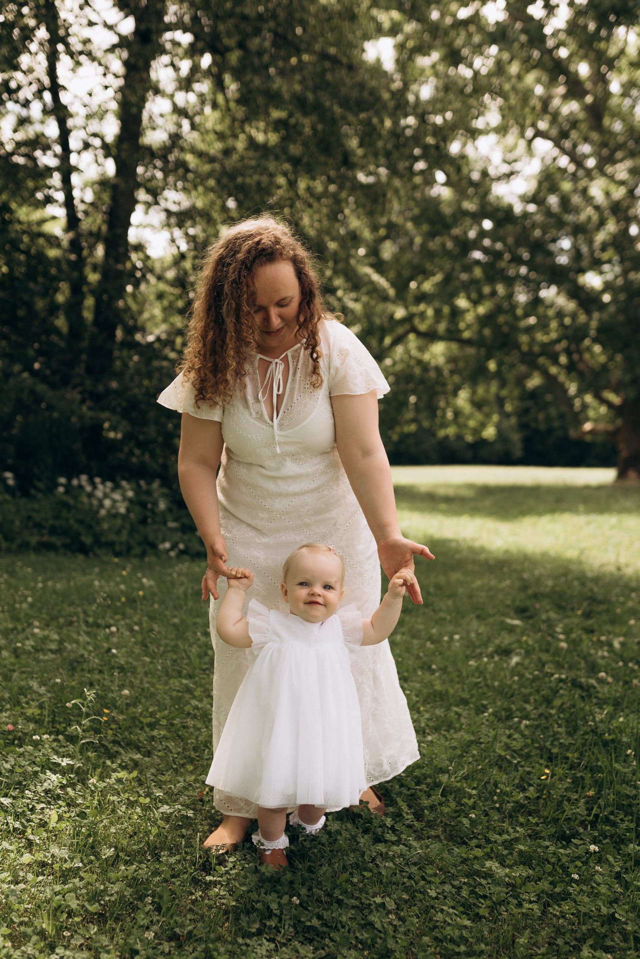 Eine Frau in einem weißen Kleid hält ein kleines Mädchen in einem weißen Kleid im Freien auf einer Wiese. Im Hintergrund sind Bäume und das Sonnenlicht. Das Mädchen lächelt und hält die Hände der Frau.