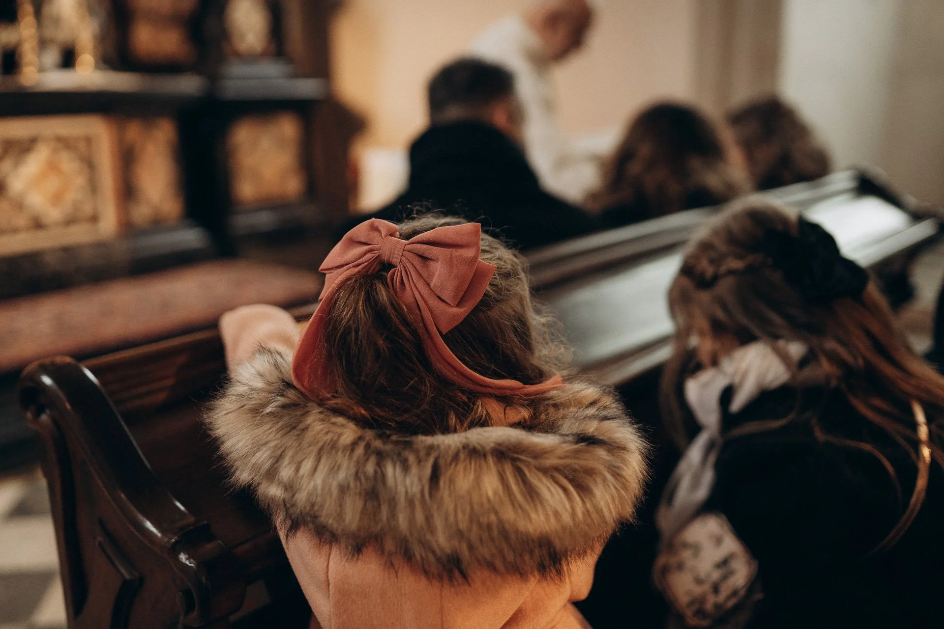 Eine Frau sitzt in einem gemütlichen Raum mit Fütterungskreide, trägt einen pelzgefütterten Mantel und einen großen rosa Schleifen-Haarclip, während sie andere Menschen im Hintergrund in einer Reihe sitzen sieht.