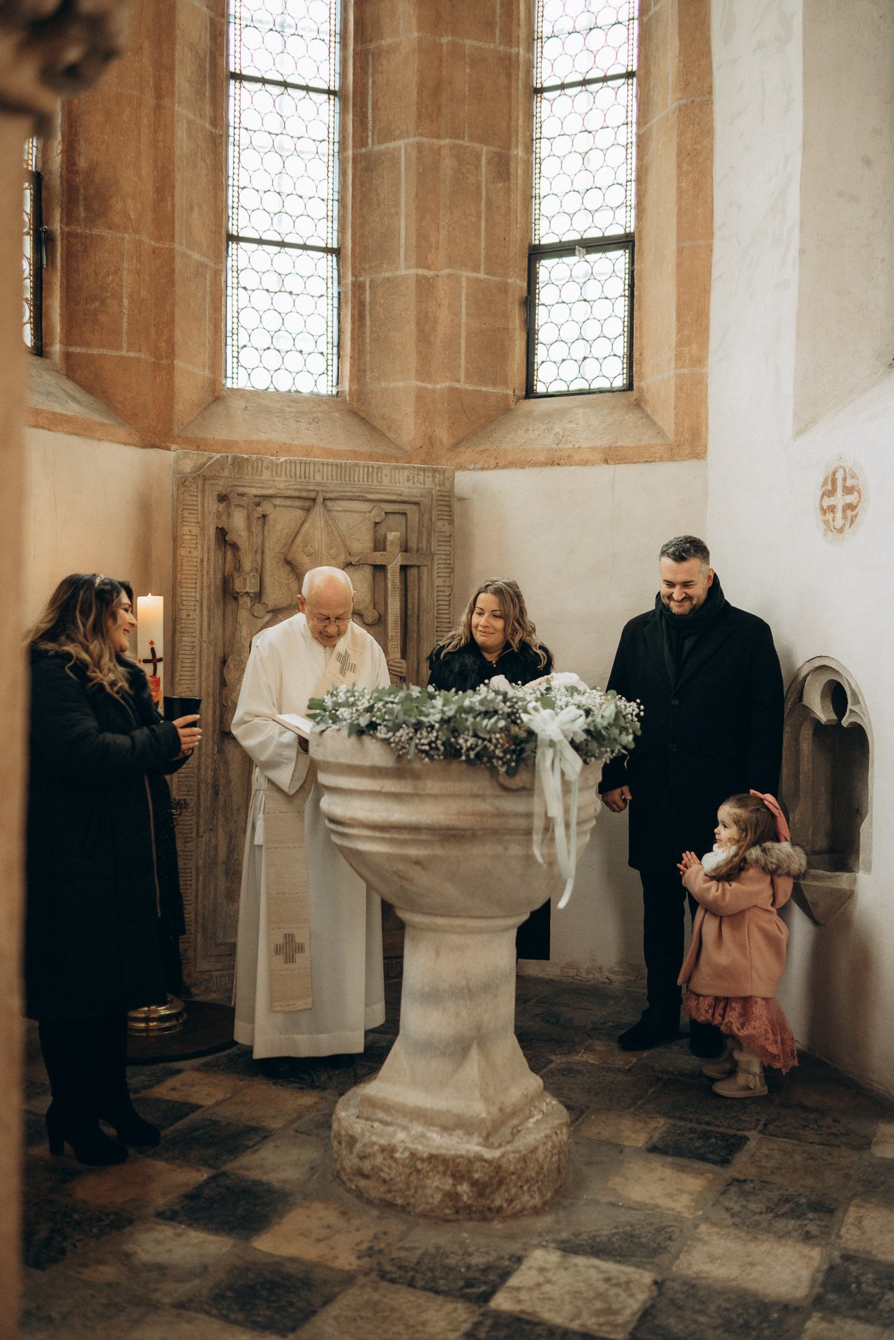 Menschen bei einer Taufe in einer Kirche, vorne ein Taufbecken, im Hintergrund große Fenster, Wand mit niedriger Mauer und Skulptur.