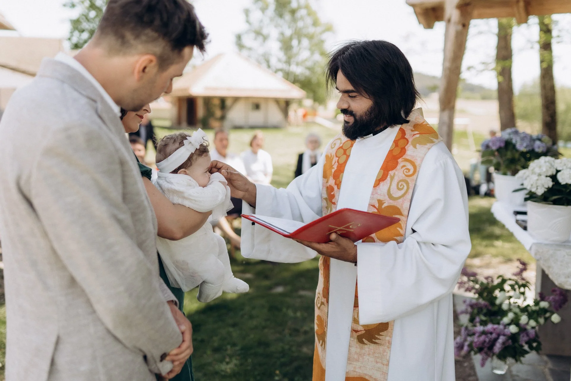 Ein Priester segnet ein kleines Mädchen bei einer Taufe im Freien, während ein Mann und eine Frau zukunftig im Vordergrund stehen.