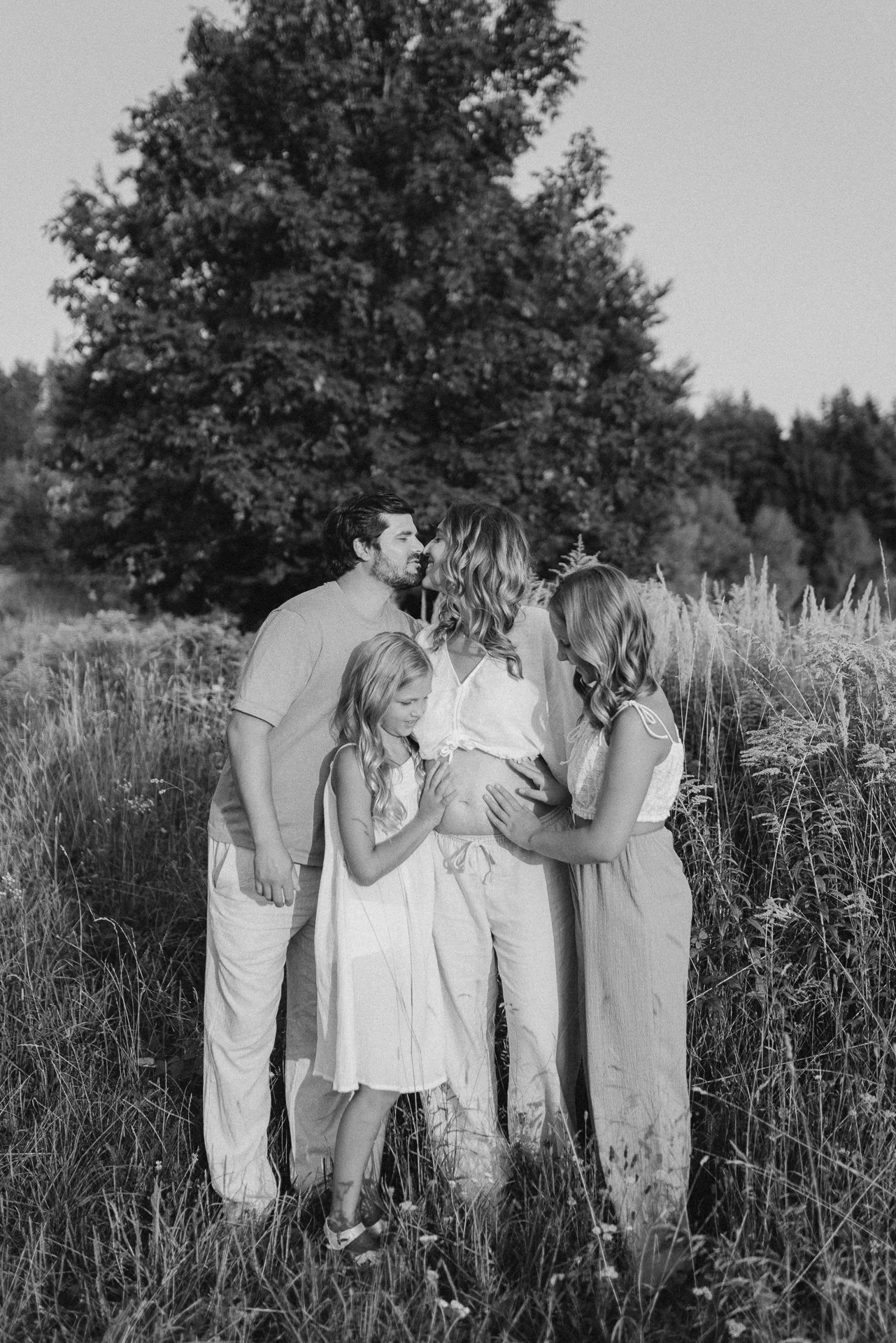 Eine Familie aus fünf Personen, darunter eine schwangere Frau, steht in einem blühenden Feld vor einem großen Baum, umarmt sich und zeigt Zuneigung in einer ländlichen Umgebung.