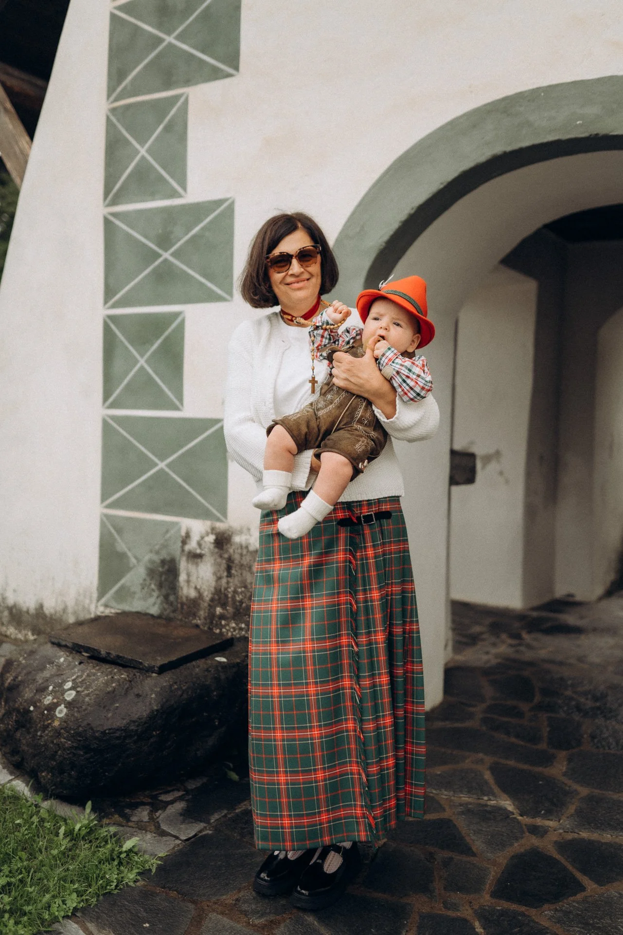Eine Frau hält ein baby in der hand vor einer weißen Mauer mit grünen geometrischen Mustern, die eine architektonische Struktur mit einem Bogen bildet. Die Frau trägt eine weiße Jacke, eine knielange Rock mit rot-grünem Karo-Muster, eine Sonnenbrille