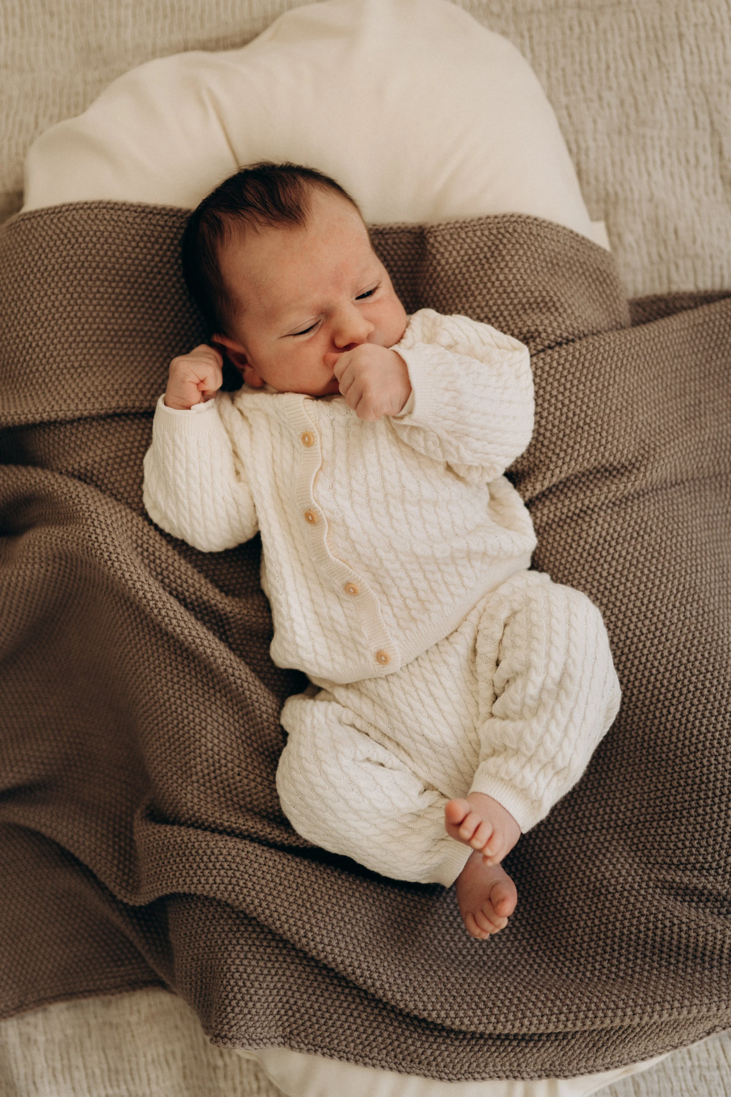 Ein kleiner Baby in einem weißen Strickanzug liegt auf einer Couch mit braunem Kissen.