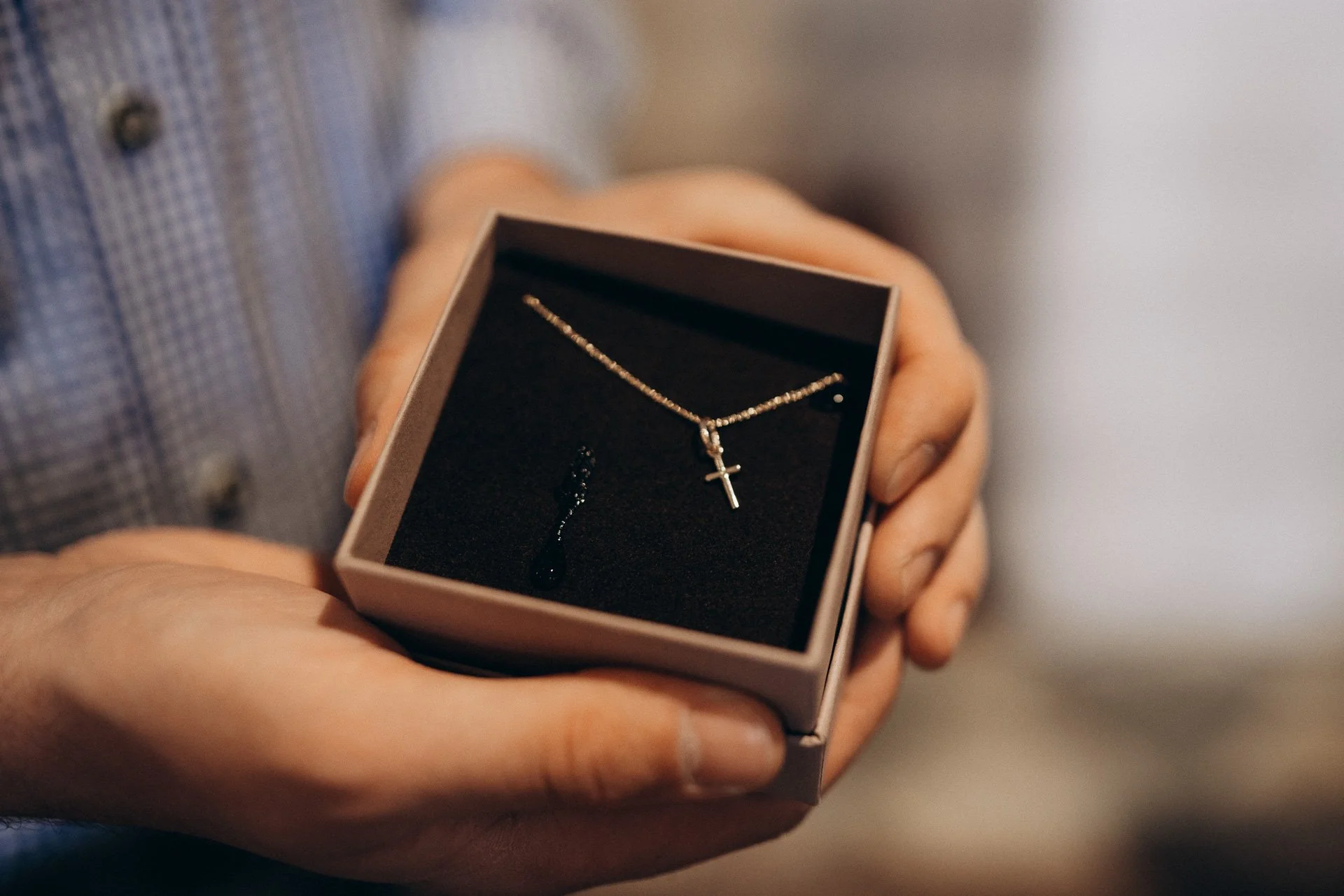 Eine Hand hält eine kleine Geschenkbox mit schwarzem Samtinnenfutter, das eine Halskette mit Kreuzanhänger und einen Ohrring enthält.