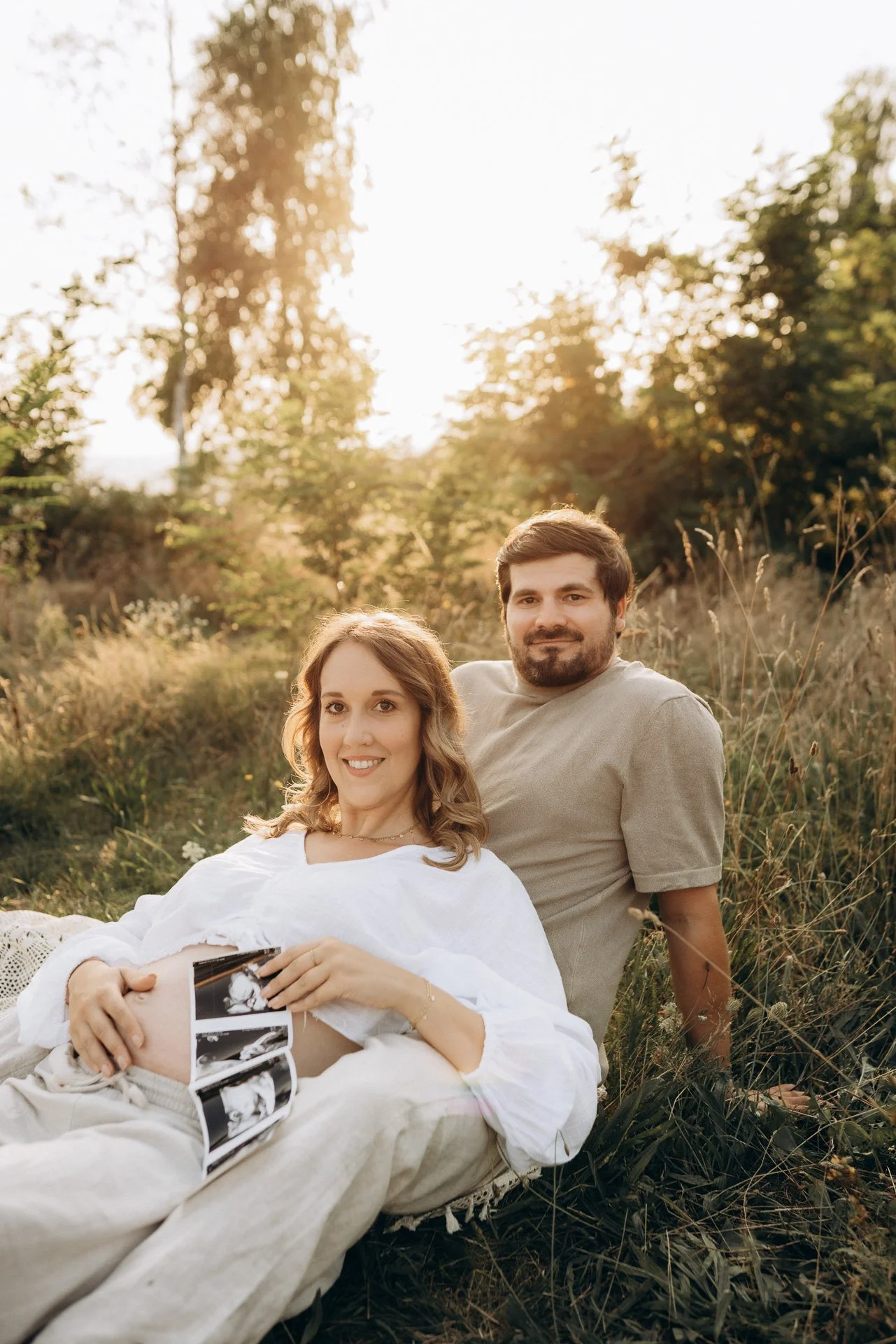 Ein glückliches Paar sitzt im Freien auf einer Wiese bei Sonnenuntergang, die Frau hält ein Ultraschallbild in den Händen und zeigt ihren Babybauch.