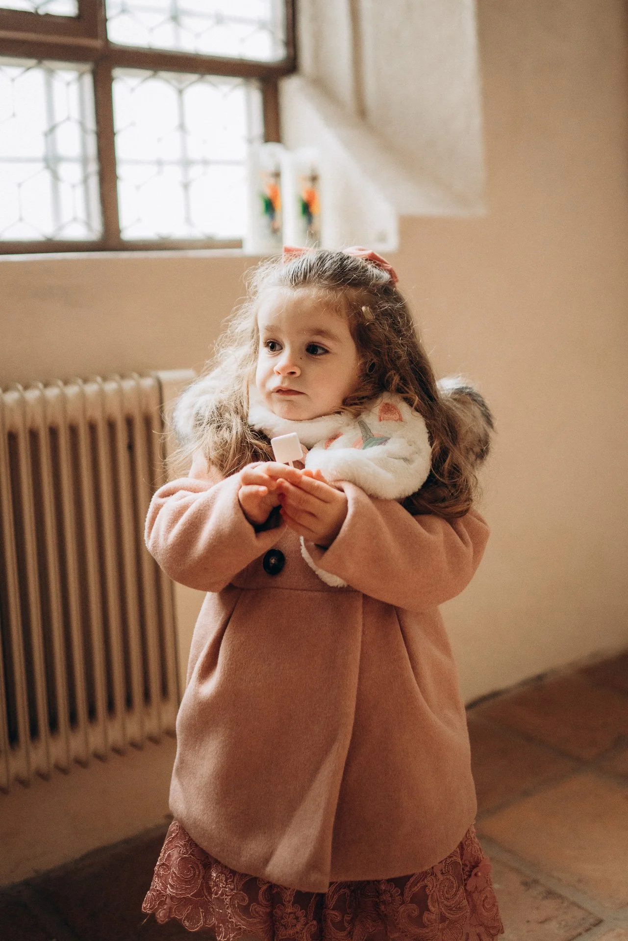 Ein junges Mädchen mit braunem, lockigem Haar, das in einem warmen Raum steht, trägt einen beige Mantel und einen rosafarbenen Rock. Sie hält eine kleine Pflaster in den Händen.