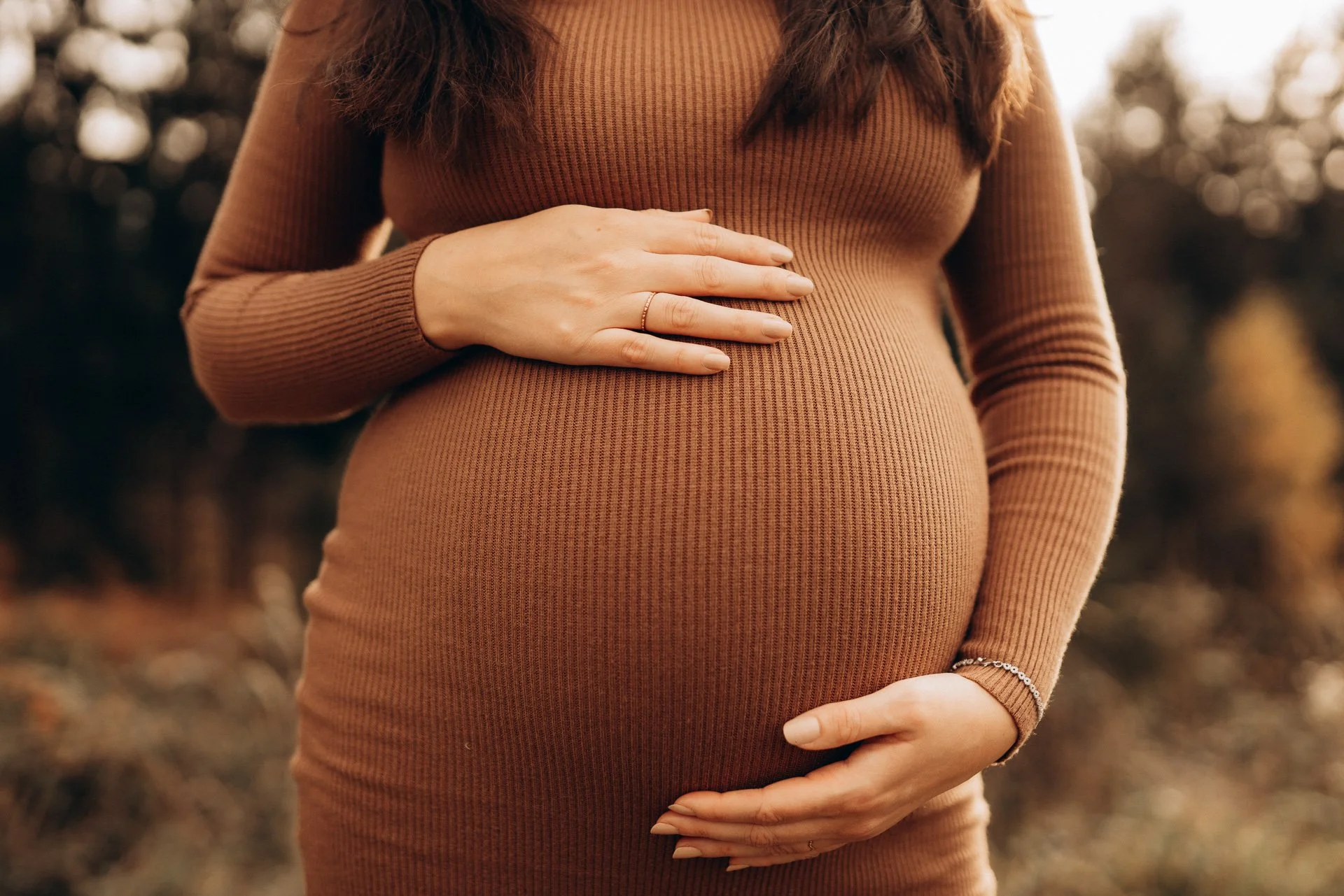 Eine schwangere Frau trägt einen engen braunen Pullover und hält ihre rechte Hand auf den Bauch, während sie im Freien steht.