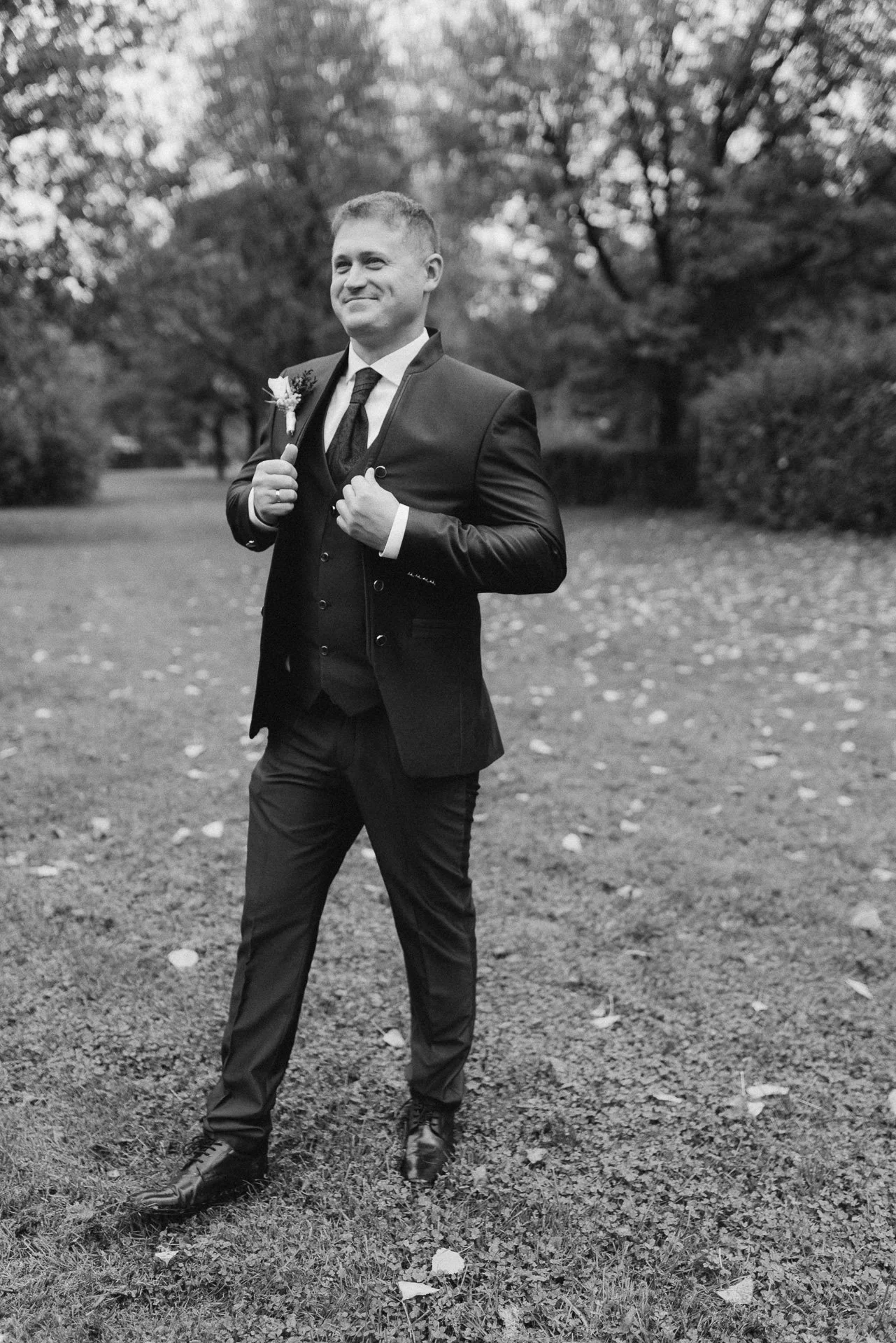 Ein Mann in einem schwarzen Anzug mit Fliege steht im Freien auf einer Wiese mit Bäumen im Hintergrund. Er lächelt und hält einen Blumenstrauß an seiner Jacke während eines Hochzeitsfotoshootings in Klagenfurt, Kärnten.