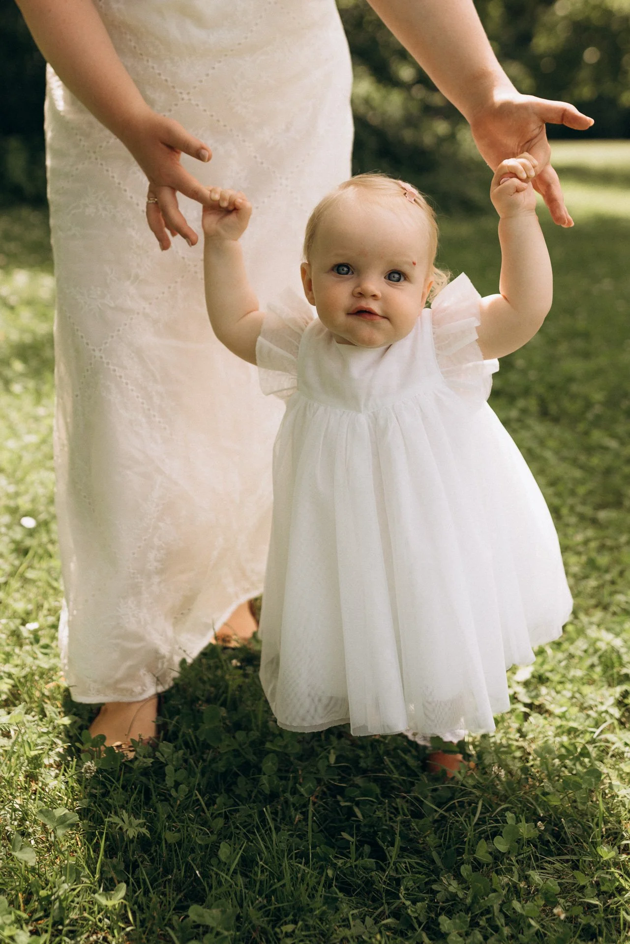 Ein kleines Mädchen in einem weißen Kleid hält die Hand einer erwachsenen Person im Freien. Die Erwachsenen trägt ein langes, cremefarbenes Kleid, und sie stehen auf einer Wiese mit grünen Pflanzen. Es ist wahrscheinlich ein Fest oder eine besondere 