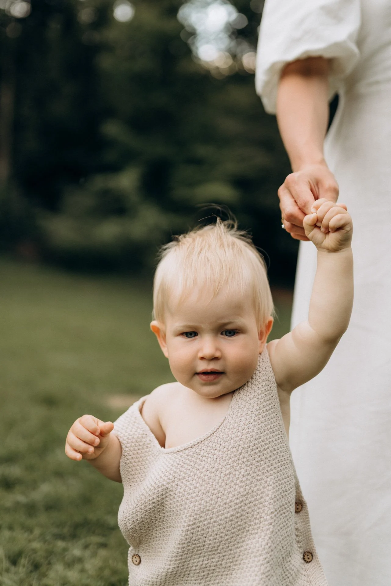 Ein kleines blondes Mädchen in einem beige Kleid, das draußen im Park gehalten wird, mit jemandem, der hinter ihm steht.