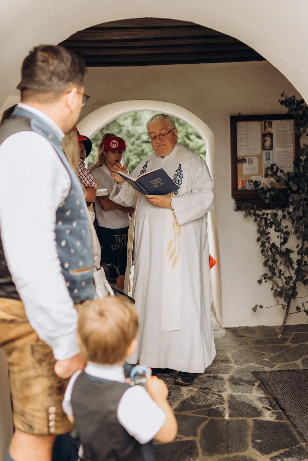 Ein Priester liest aus einem Buch vor, während eine Gruppe von Kindern und Jugendlichen aufmerksam zuhört. Die Szene ist in einem Raum mit weißer Wand und einer quadratischen Steinboden. Es scheint sich um eine Veranstaltung oder Zeremonie zu handeln