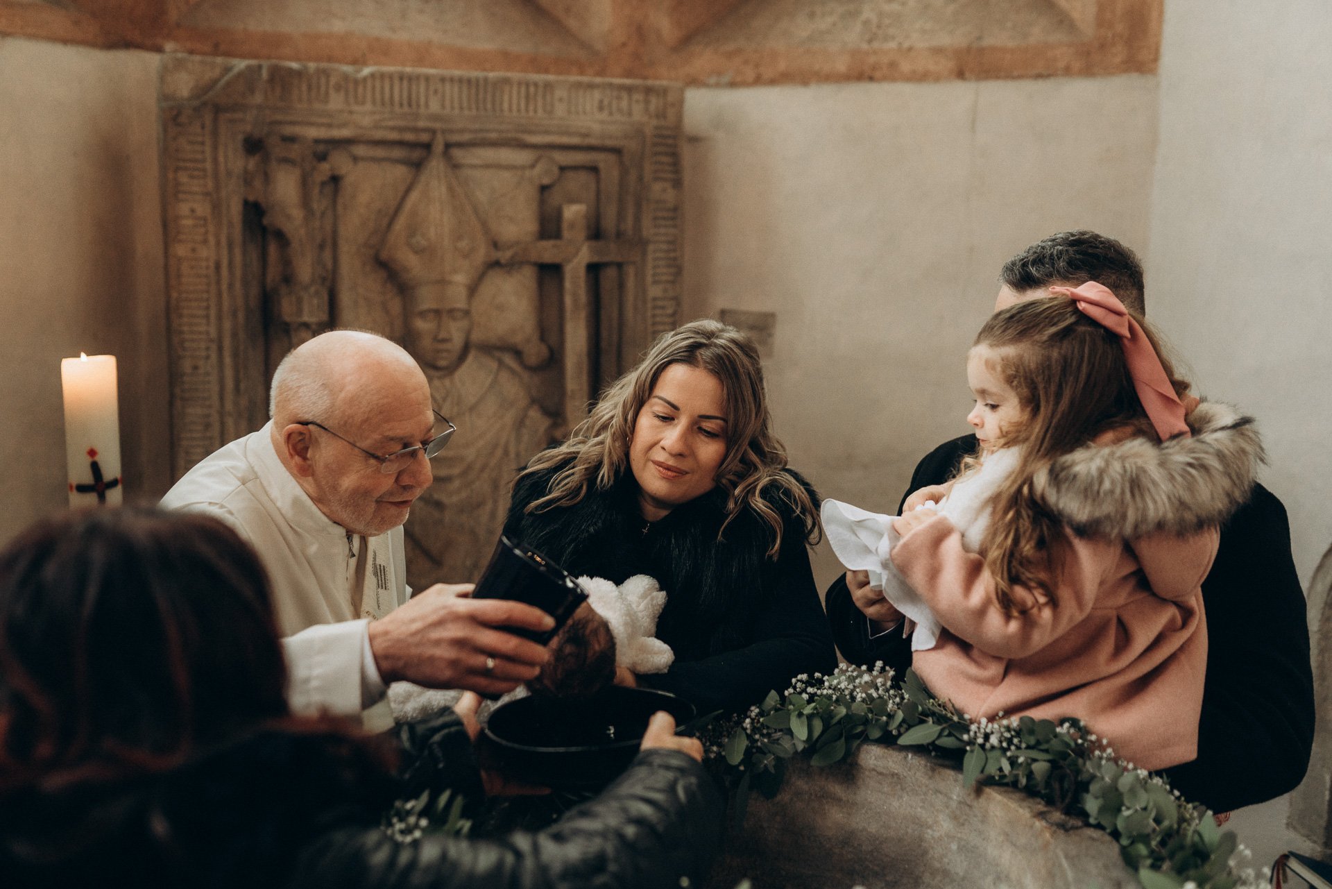 Familie bei einer Taufe in einer Kirche, während ein Priester die Taufe vollzieht, mit Kindern und Erwachsenen, die anwesend sind.