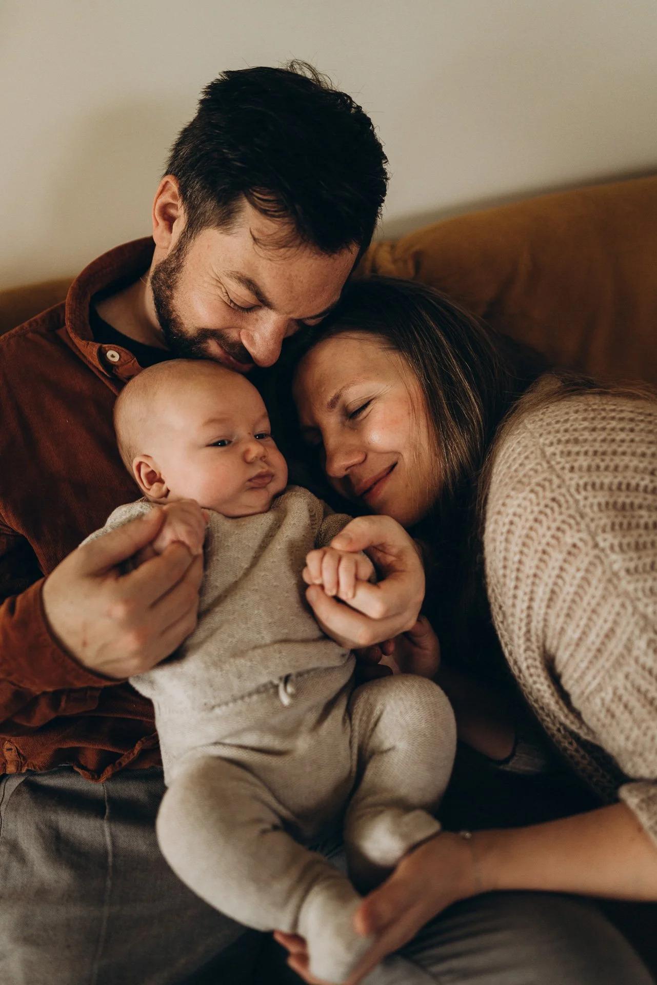 Familie mit Mutter, Vater und Baby, die liebevoll zusammen auf einem Sofa kuscheln.