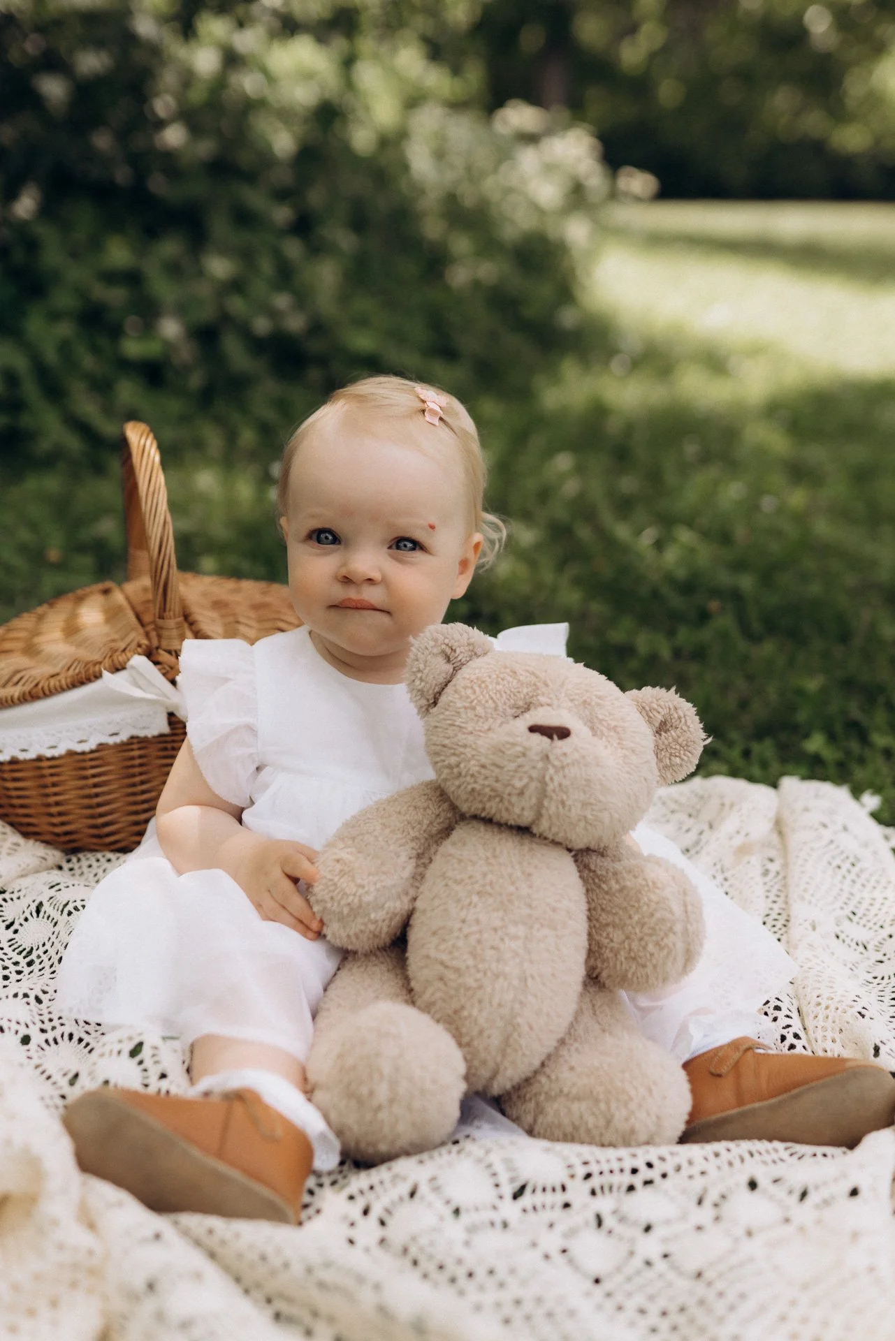 Ein kleines Mädchen in Weiß sitzt auf einer Decke im Grünen, hält einen braunen Kuschelteddybären und hat einen Korb im Hintergrund.