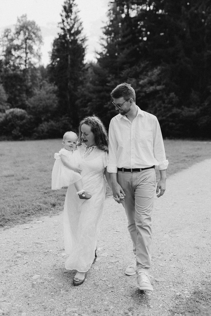 Ein glückliches Familienbild, in dem eine Frau eine kleine Tochter trägt, während ein Mann und die Frau Hand in Hand auf einem Pfad in einem Wald spazieren.
