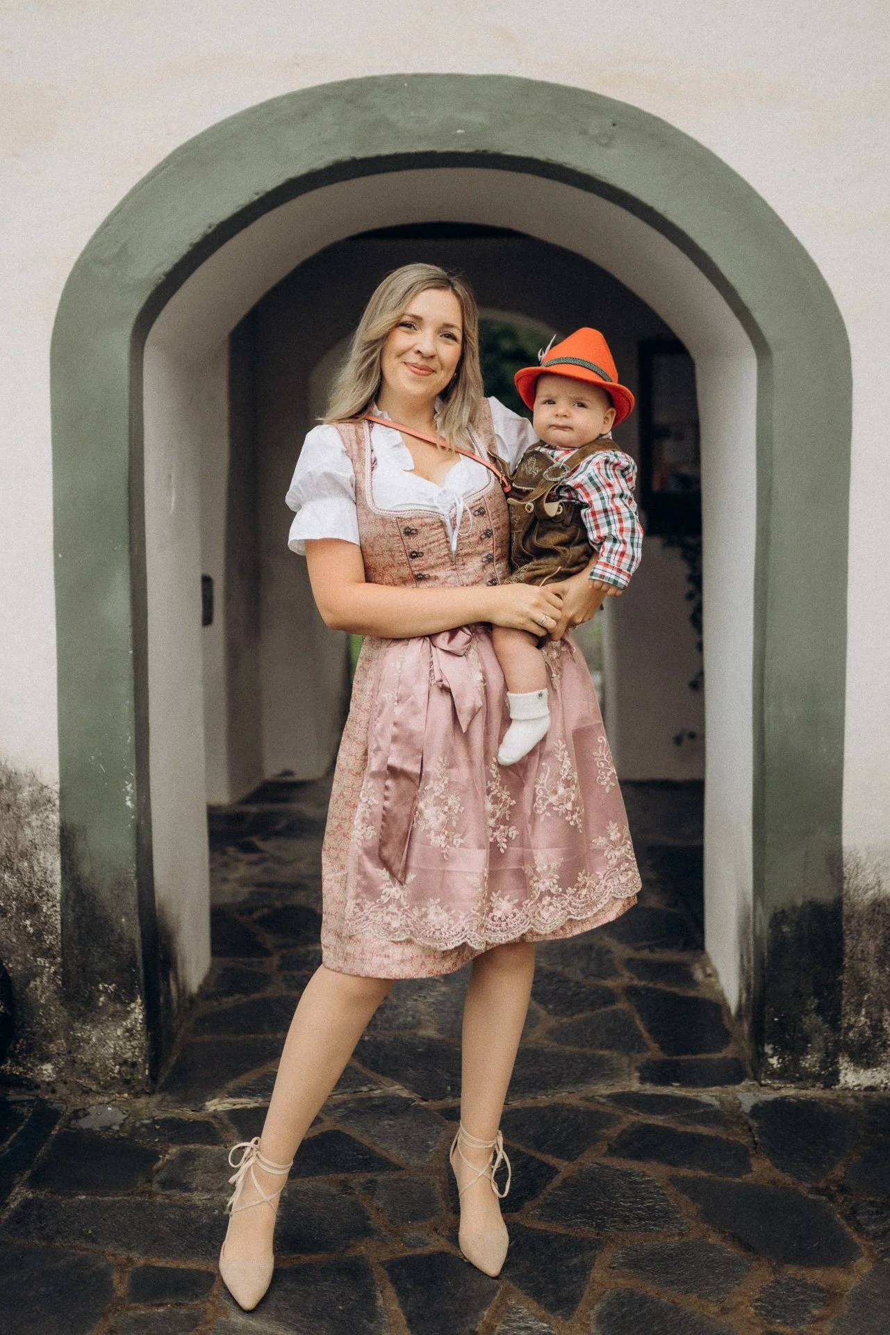 Eine Frau in traditioneller Tracht hält ein Baby, das ebenfalls in Tracht gekleidet ist, vor einer Arkade.