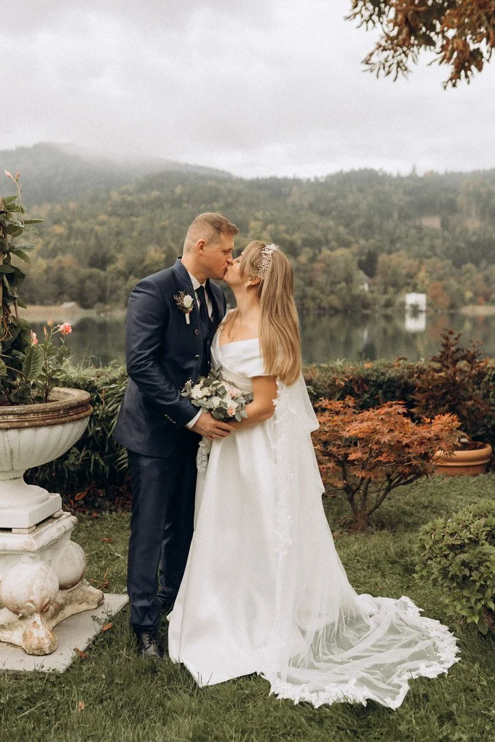 Ein Brautpaar bei ihrer Hochzeit, das sich liebevoll küsst, im Freien vor einer seenähnlichen Wasserfläche mit Berg im Hintergrund und herbstlich gefärbtem Laub.